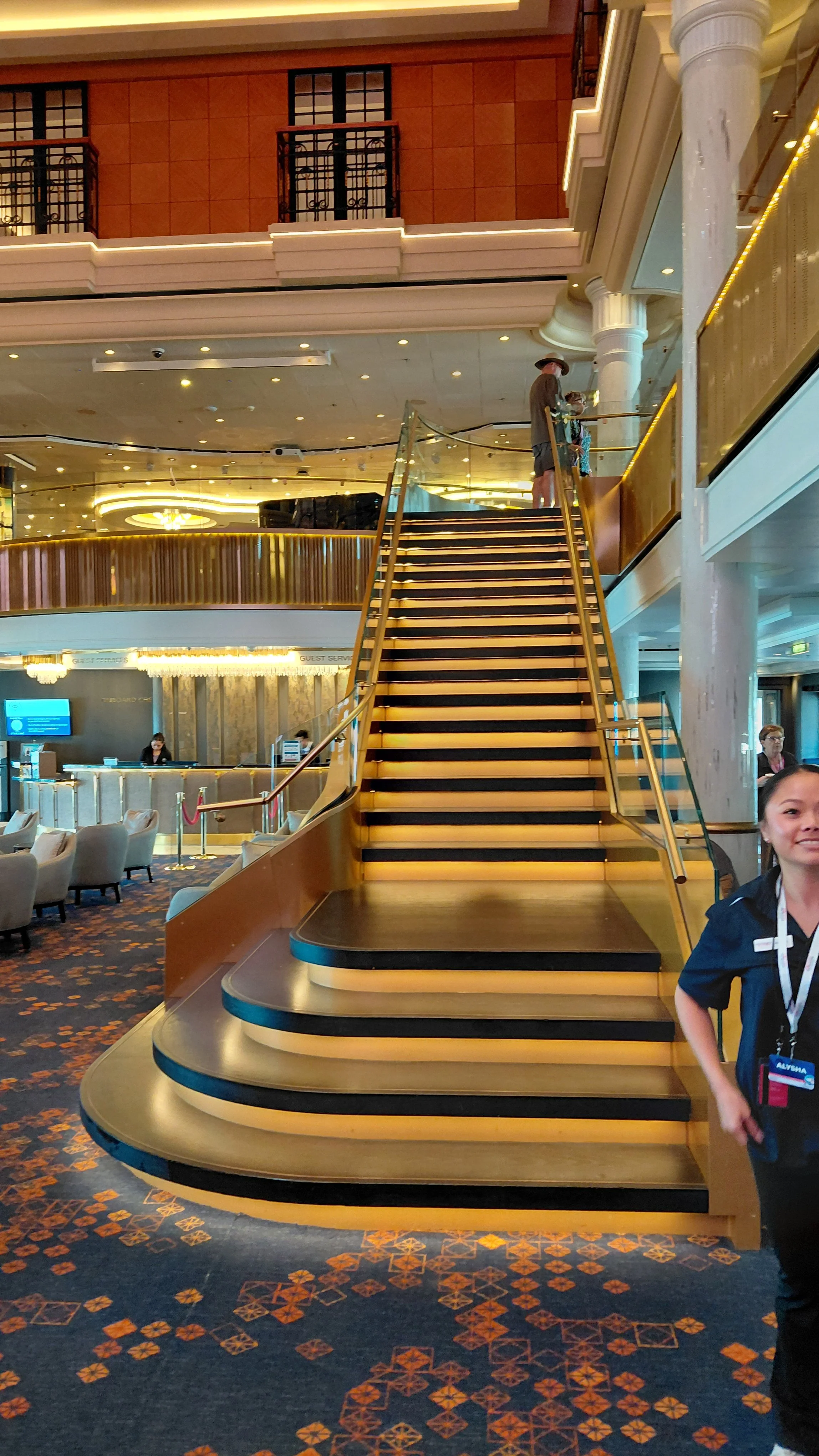 Interior of a hotel lobby with a curved staircase with gold railing and black steps, plush chairs, and a reception desk. Two people are at the top of the staircase, and a woman with a badge is in the foreground.