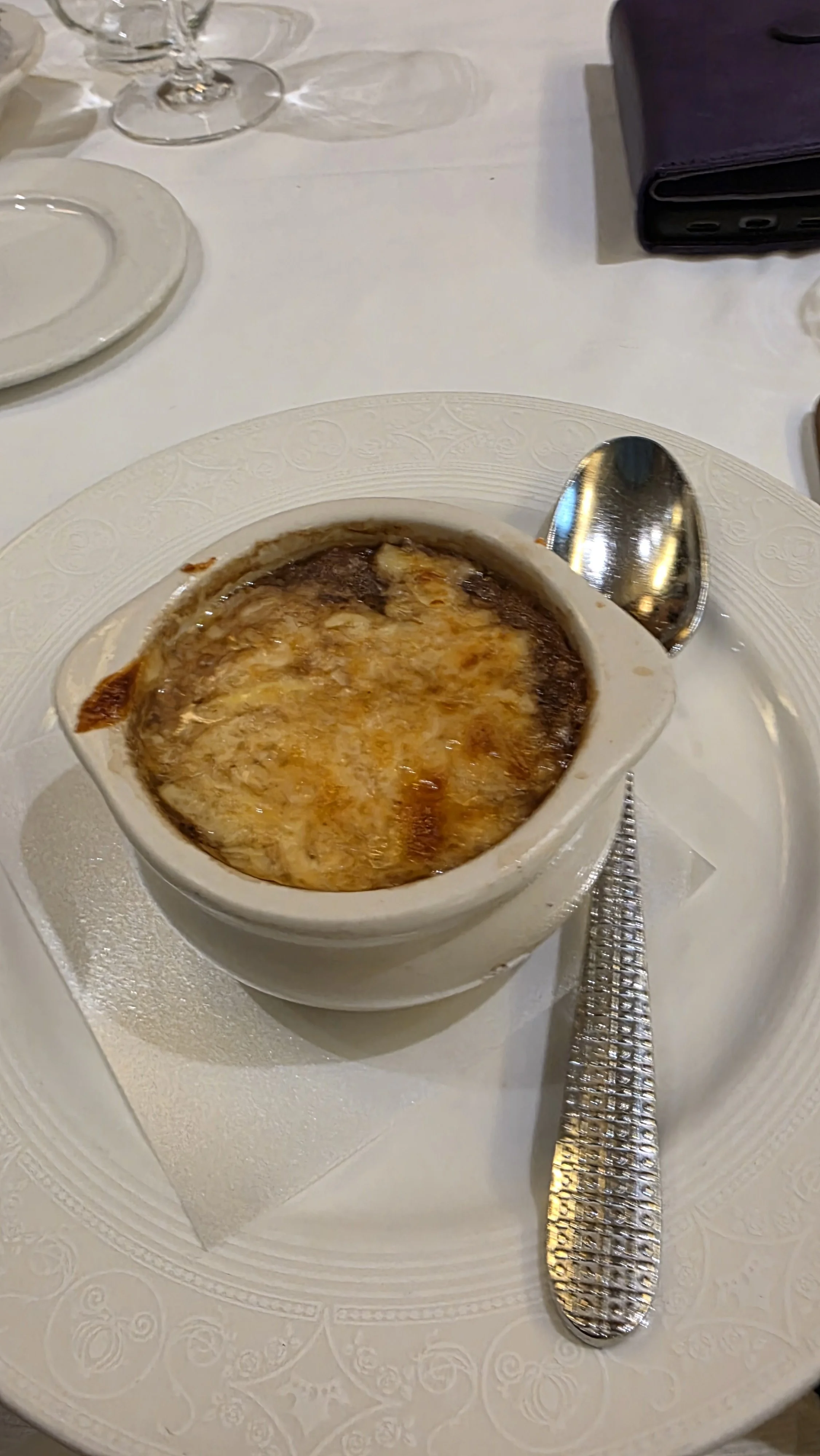 Baked French onion soup with melted cheese in a small white ramekin on a white plate with a silver spoon beside it, set on a white tablecloth with other tableware visible.