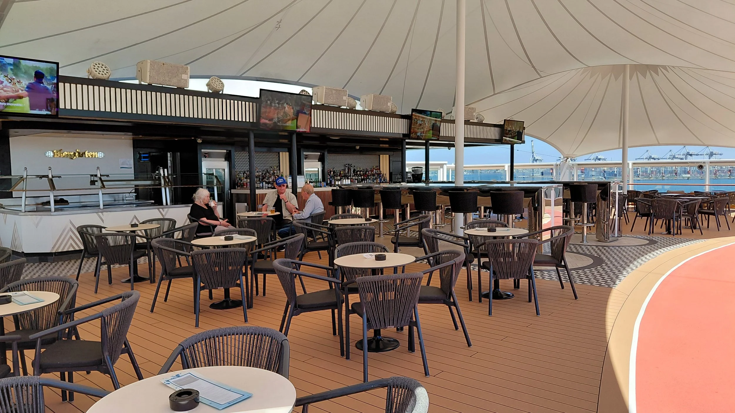 An outdoor bar and seating area on a cruise ship deck with tables, chairs, a bar counter, and a large white canopy overhead, with ships visible in the harbor in the background.