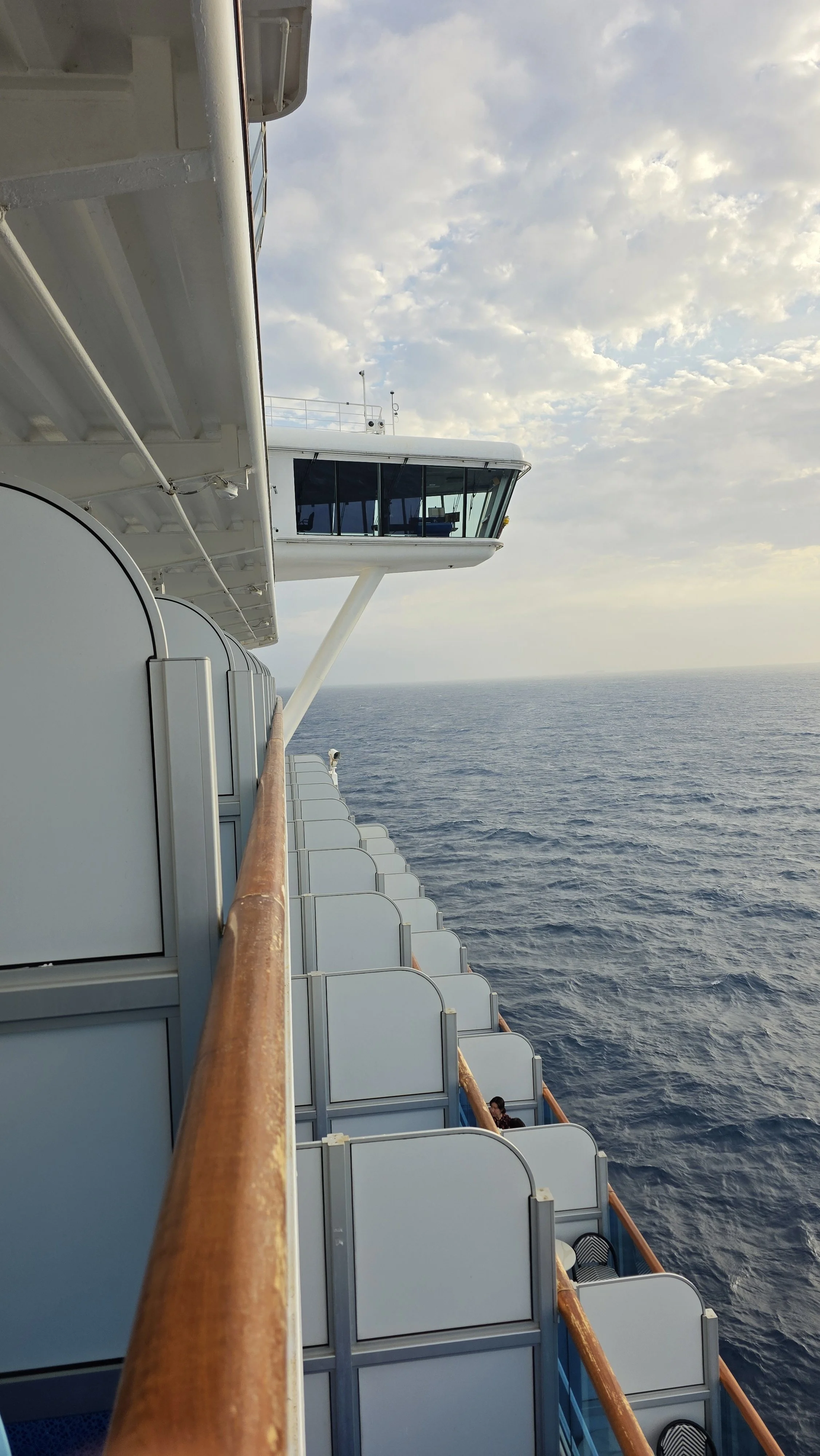 View of the side of a cruise ship with sunlit balcony cabins and the ocean in the background, taken from the ship's open deck
