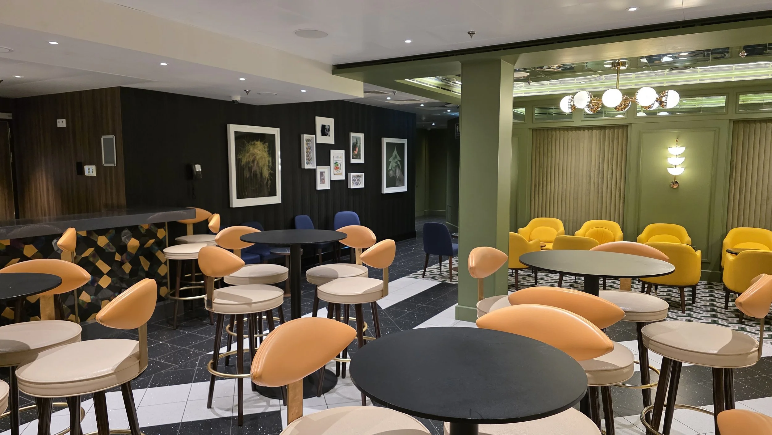 A stylish lounge area with colorful seating, including beige chairs with peach cushions, yellow armchairs, and dark blue chairs, arranged around round tables. The walls are decorated with framed artwork and modern lighting fixtures, blending dark woo
