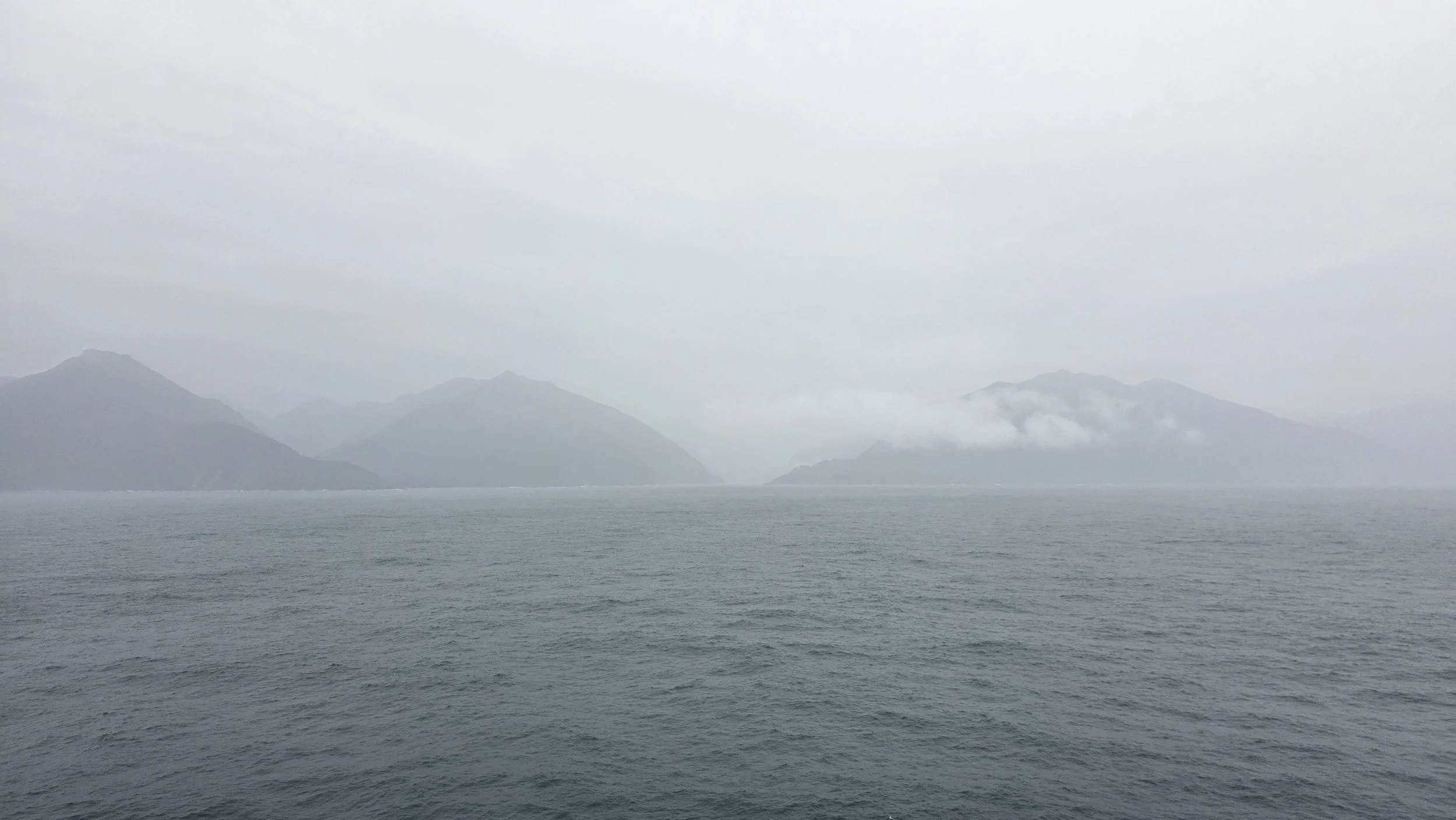A misty ocean with distant mountains partially obscured by fog and clouds.