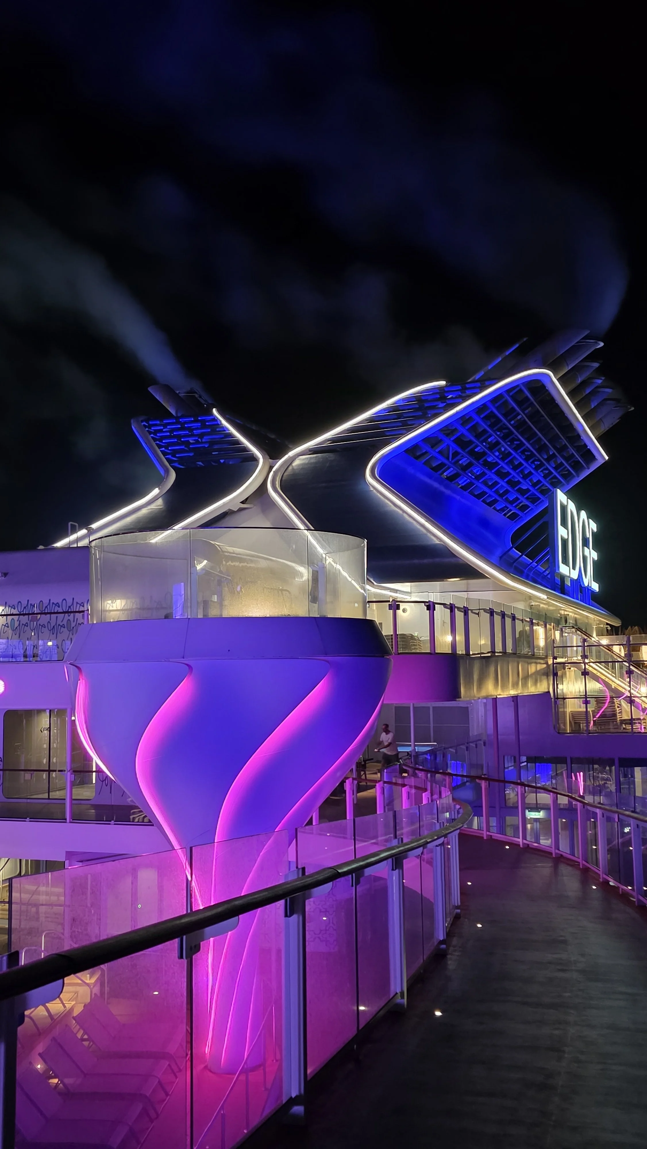 Night view of a modern, futuristic building with glowing blue and pink neon lights, labeled 'EDGE', featuring curved architectural design and glass railings.