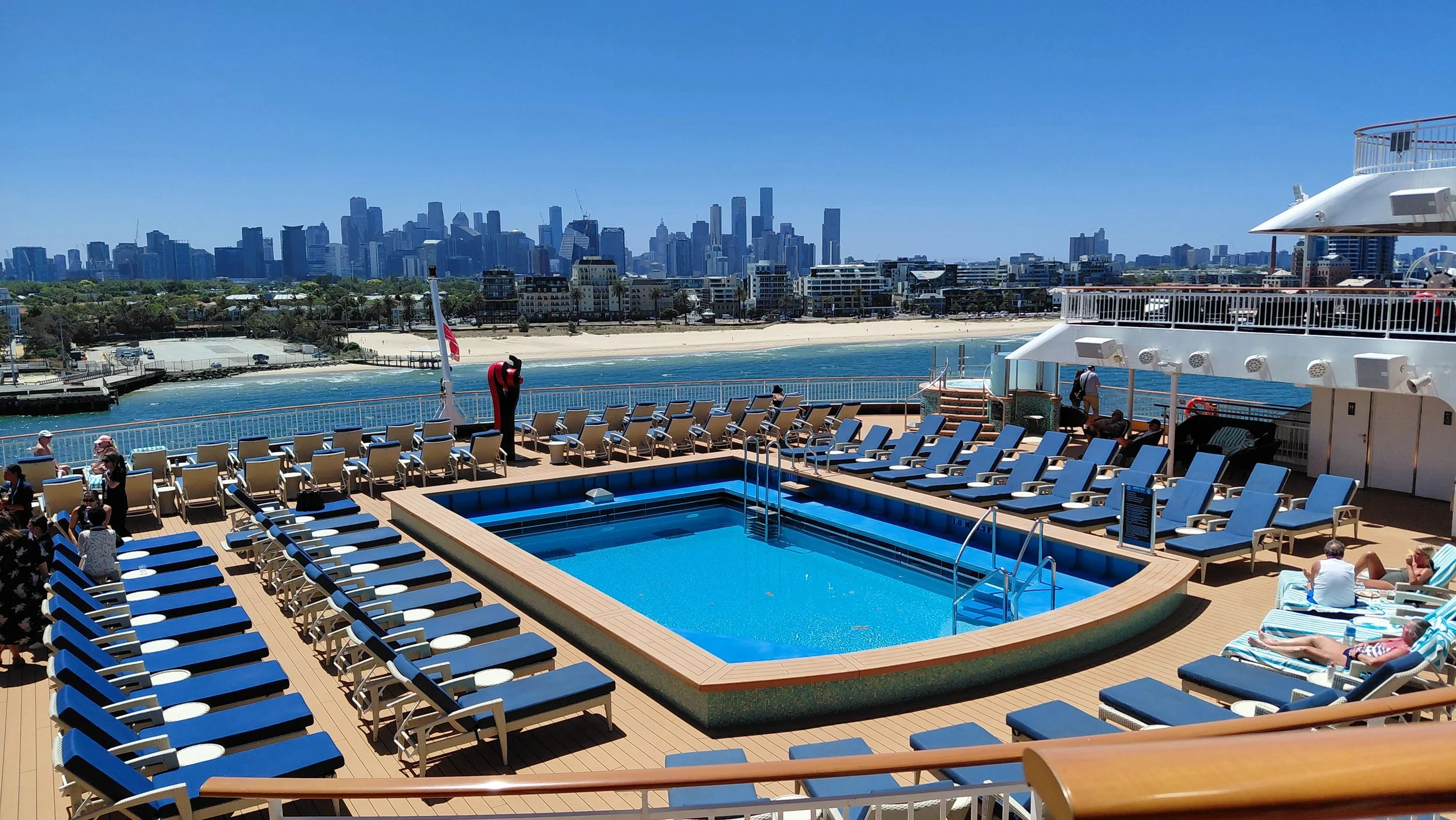 View of a cruise ship deck with a pool surrounded by lounge chairs, some people sunbathing and others walking, with a city skyline and ocean in the background.