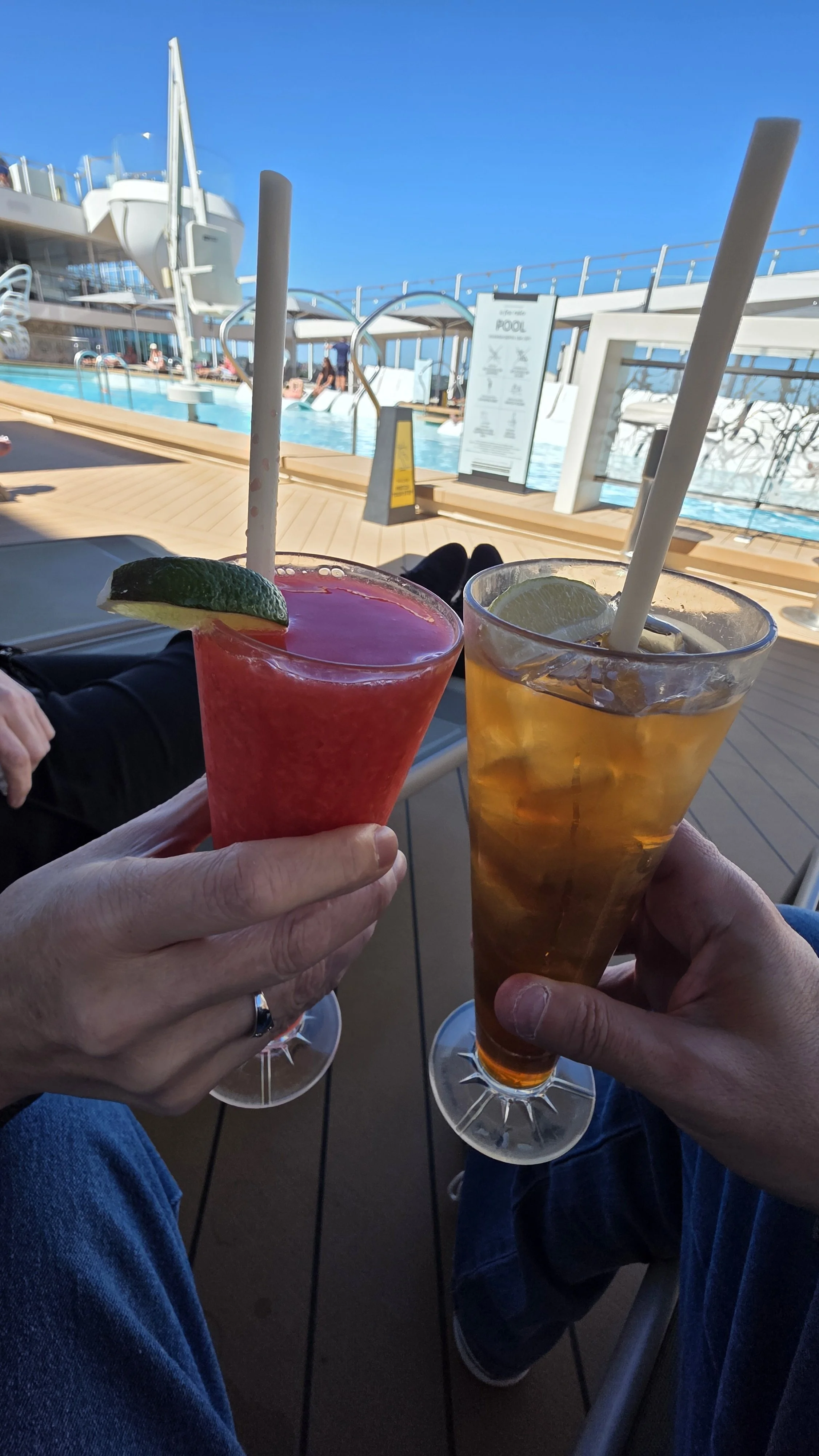 Two drinks, one pink with lime and one amber with lemon, being held up by two people by a poolside with a cruise ship in the background.