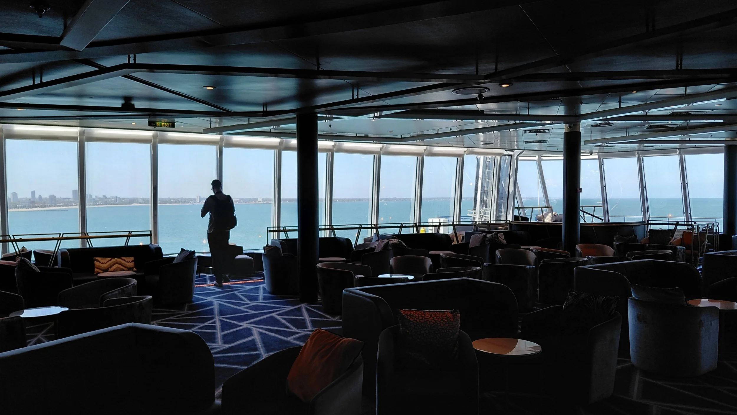 Interior of an airport lounge with large windows overlooking the ocean, a person standing near the window, and multiple chairs and sofas arranged throughout the space.