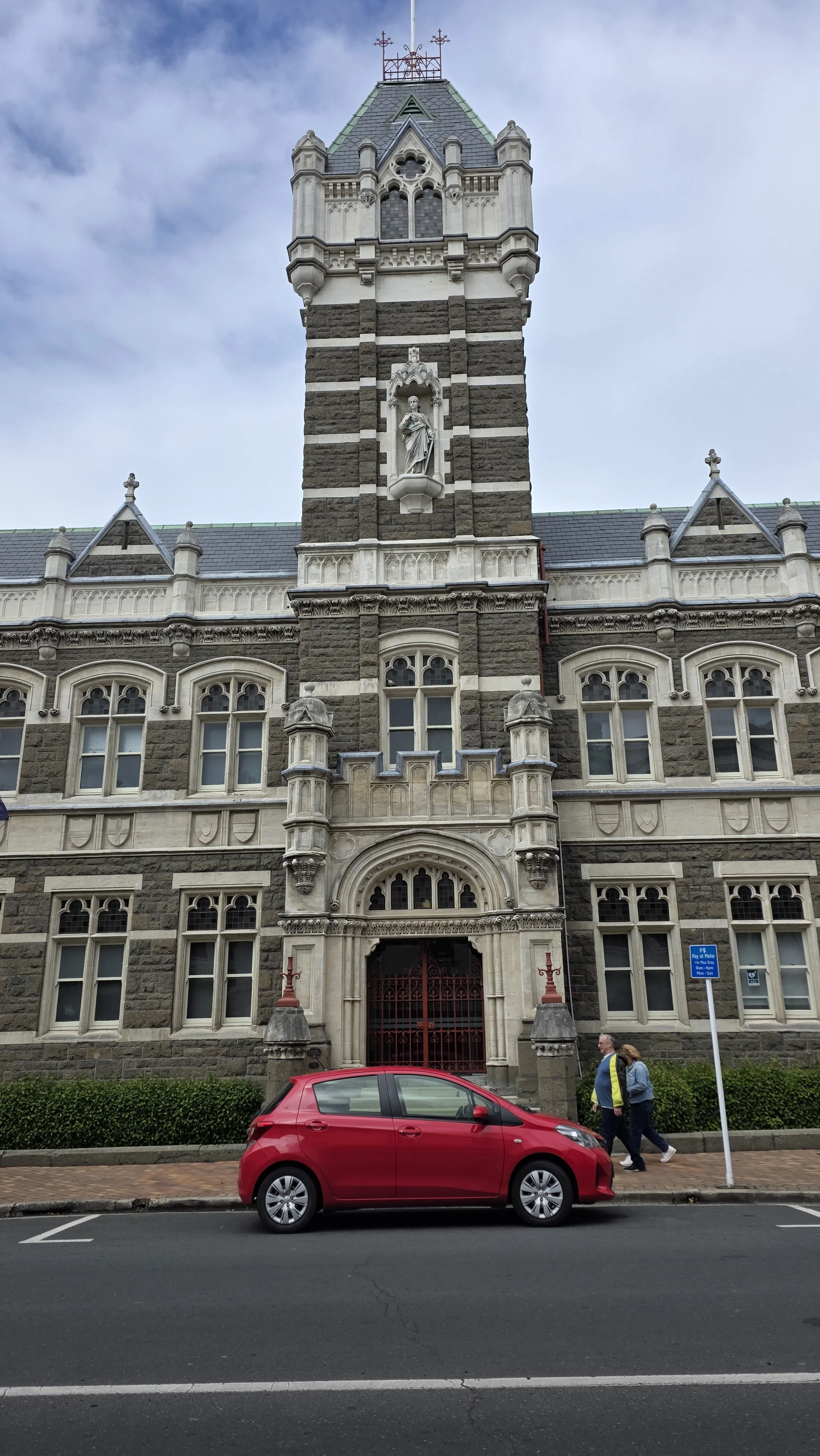 A historic stone building with Gothic architecture featuring arched windows, decorative stonework, and a tower with a statue. A red car is parked in front, and two people are walking on the sidewalk.