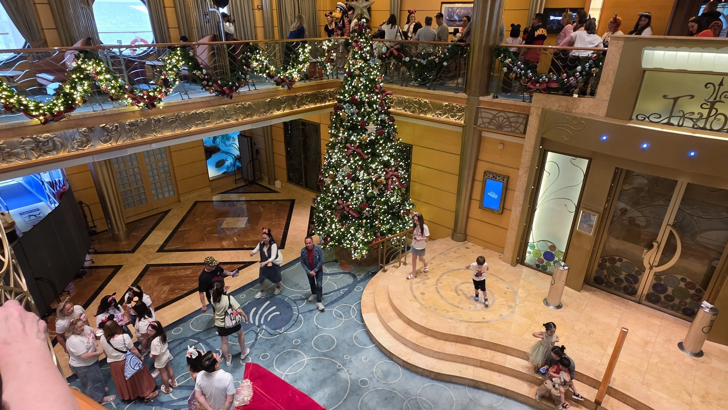 A large decorated Christmas tree with red bows, ornaments, and lights inside a spacious, elegant hotel lobby. People, including children and adults, are gathered around, some in line, some seated on steps, and some walking. The upper level has holida