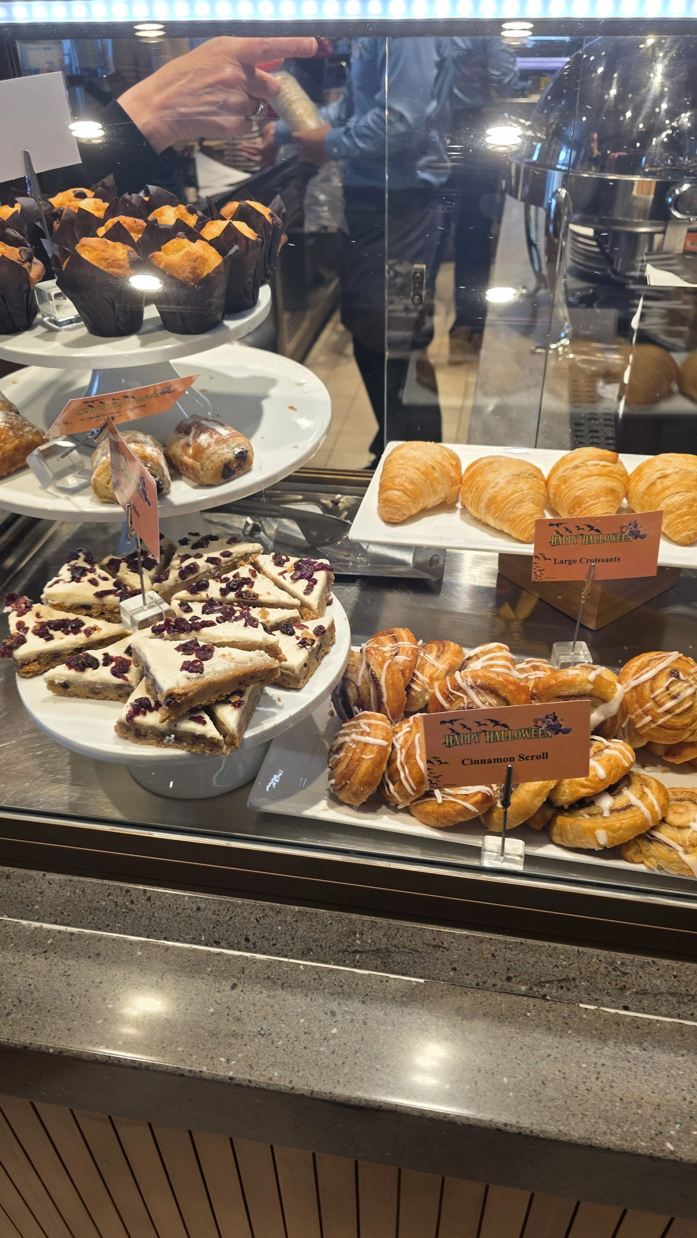 Display case with assorted baked goods including croissants, cinnamon scrolls, berry-topped cheesecakes, and muffins at a bakery or cafe.