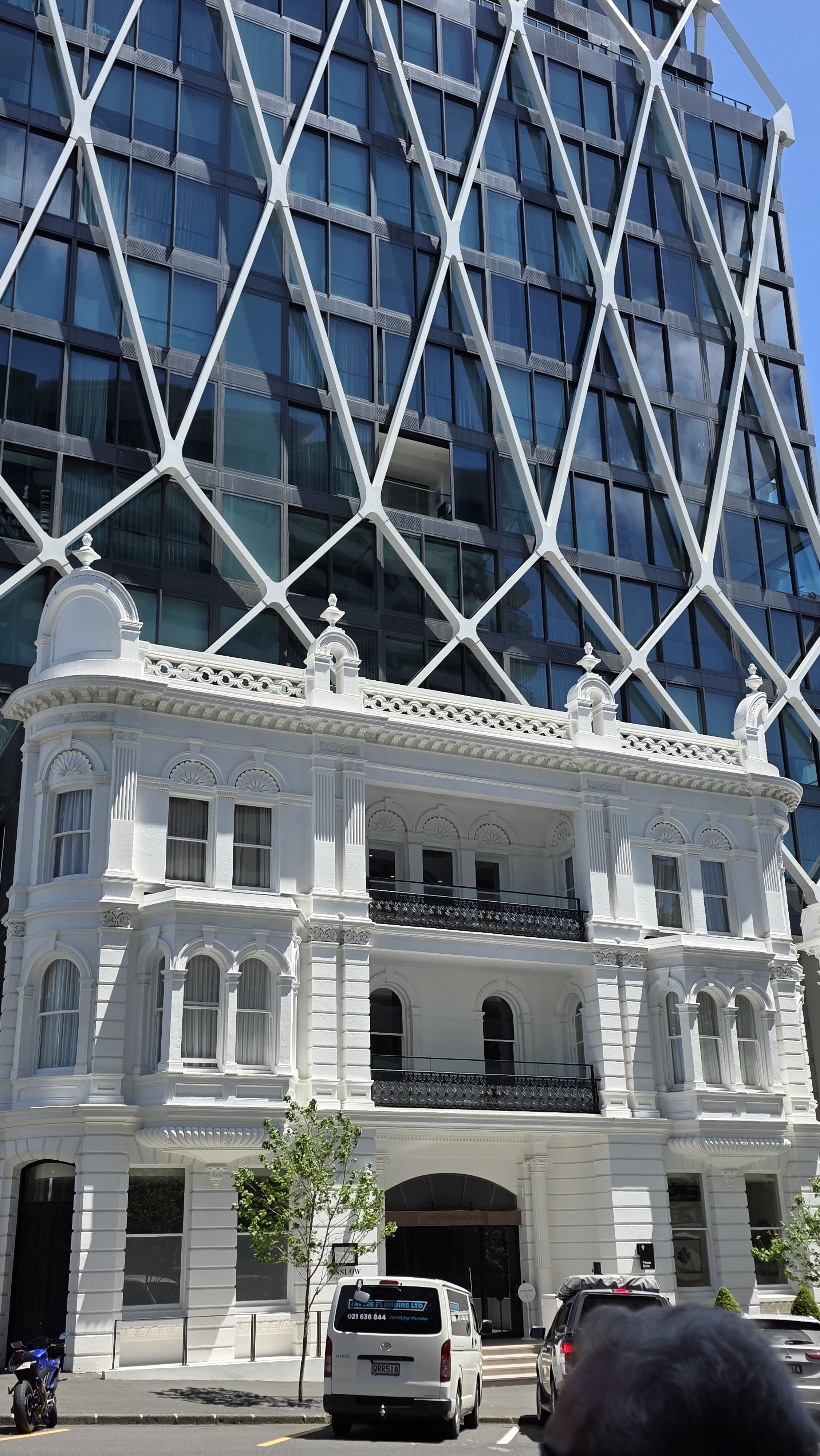 A modern high-rise glass building with a white Victorian-style house facade at the bottom, parked cars and small trees in front.