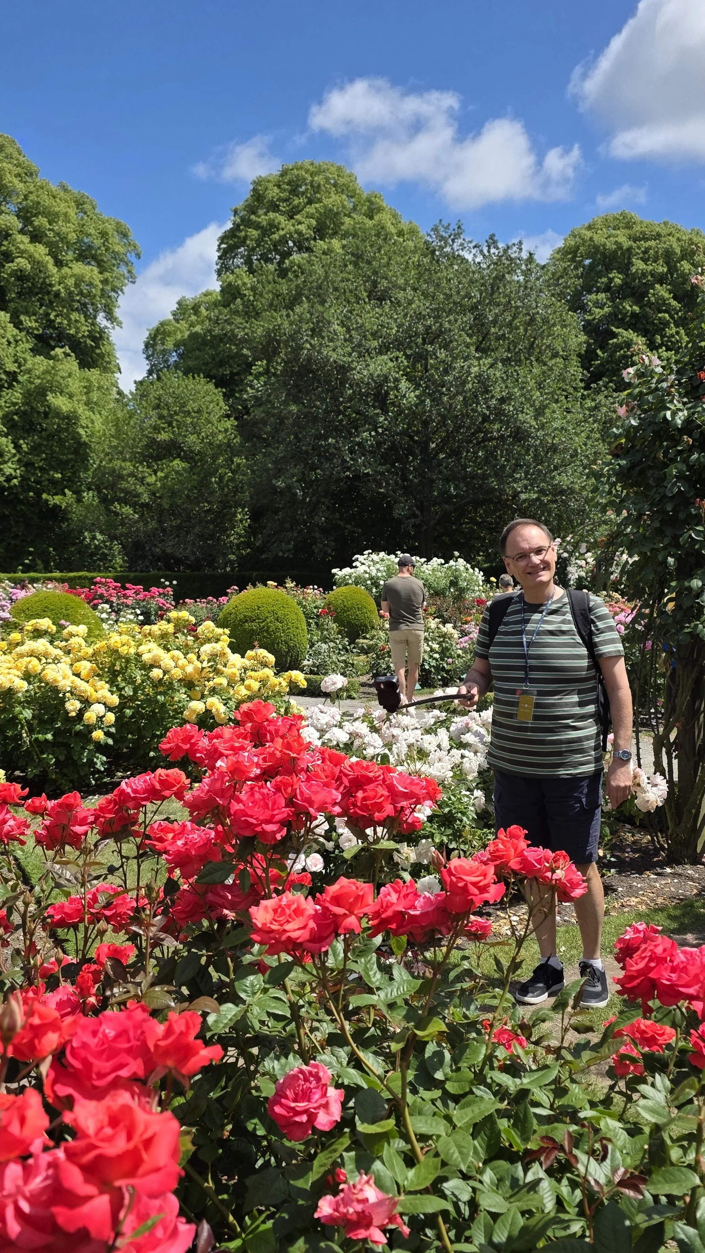 Person smiling and holding a camera in a colorful flower garden on a sunny day with trees and blue sky in the background.