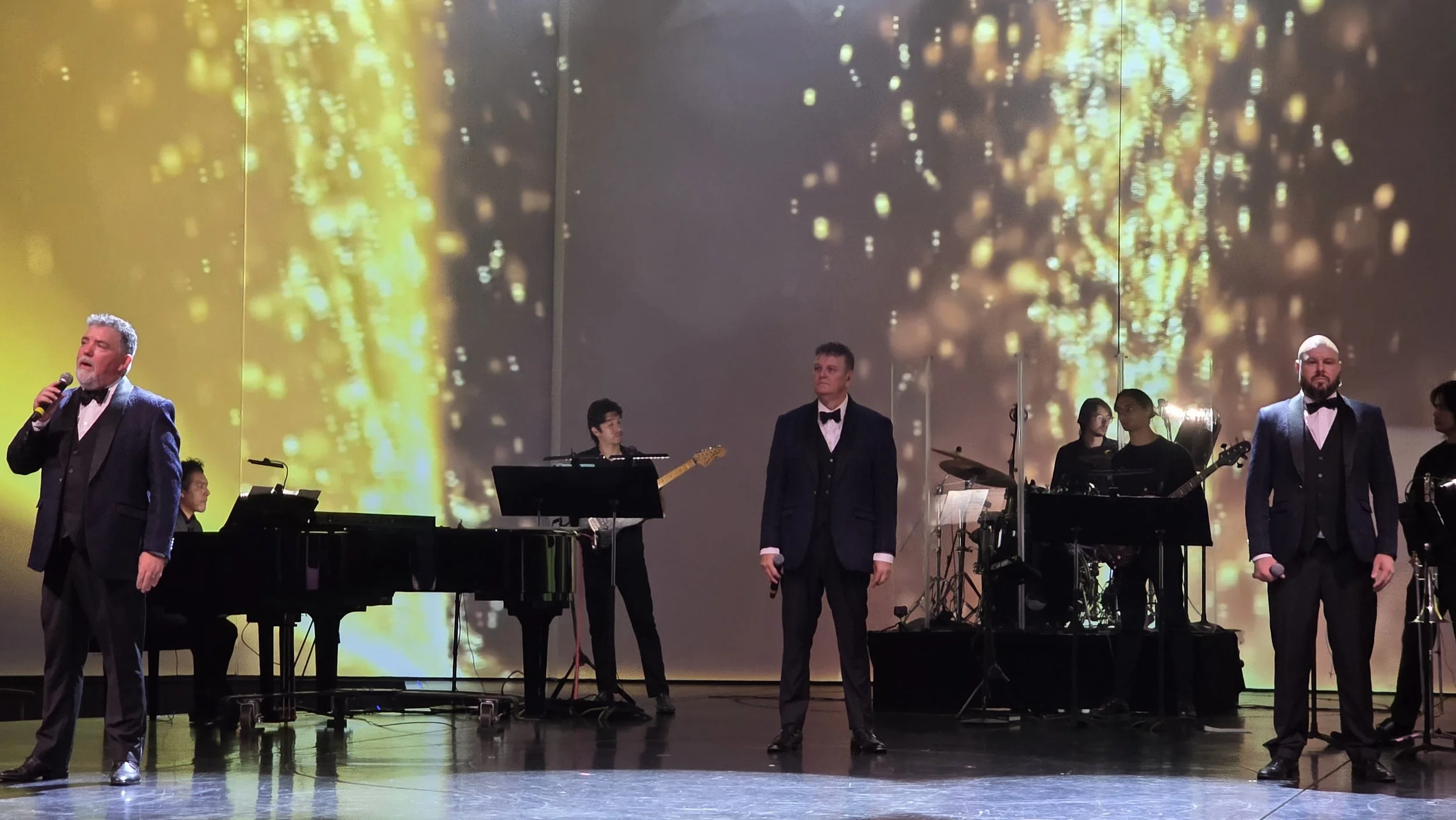 Performers on stage, with four men singing and a band with members playing piano, guitar, drums, and keyboard in the background, against a large digital screen displaying a golden, glittering pattern.