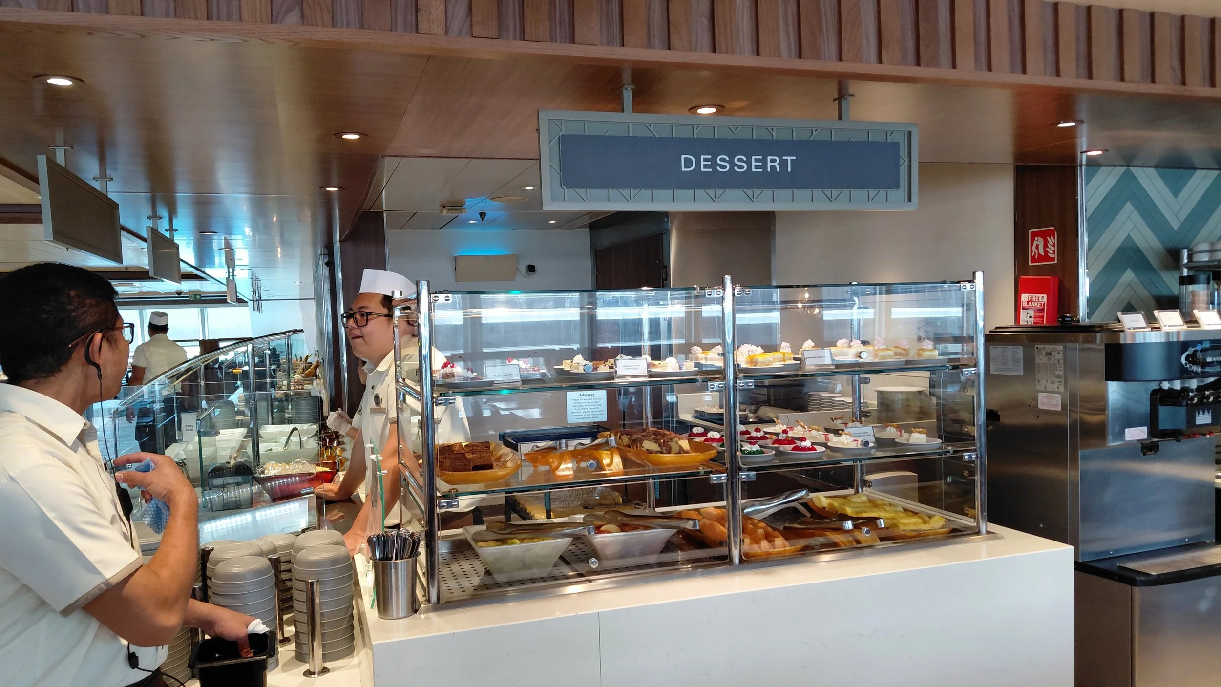A dessert buffet in a restaurant with a glass display case filled with various cakes, pastries, and fruit. Two people are engaged in conversation by the counter, one wearing glasses and the other wearing a chef's hat. A sign above reads 'DESSERT'.