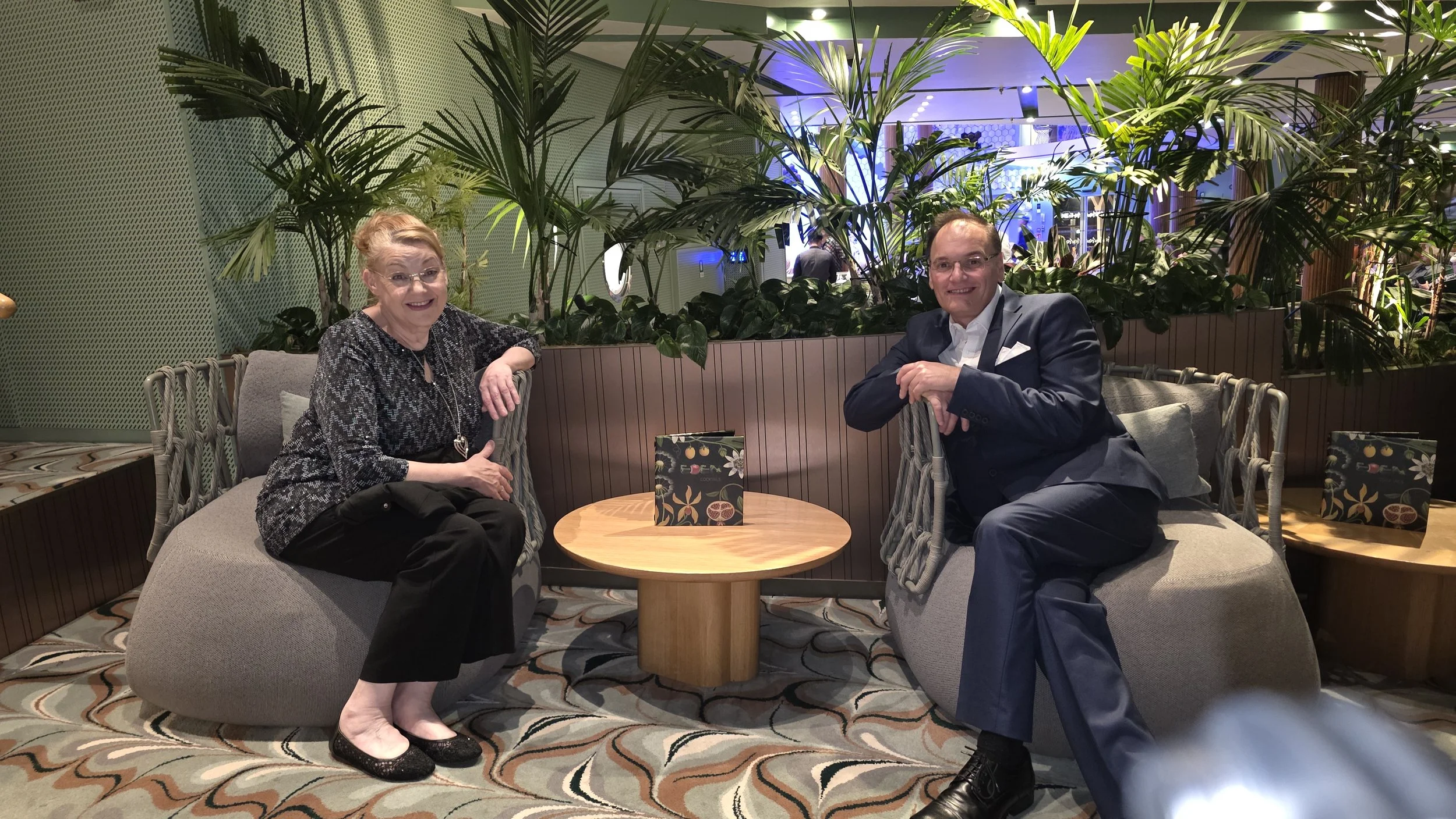 Two people sitting on modern lounge chairs in a stylish indoor setting with lush tropical plants, a wooden table with a decorative bag, and patterned carpet.