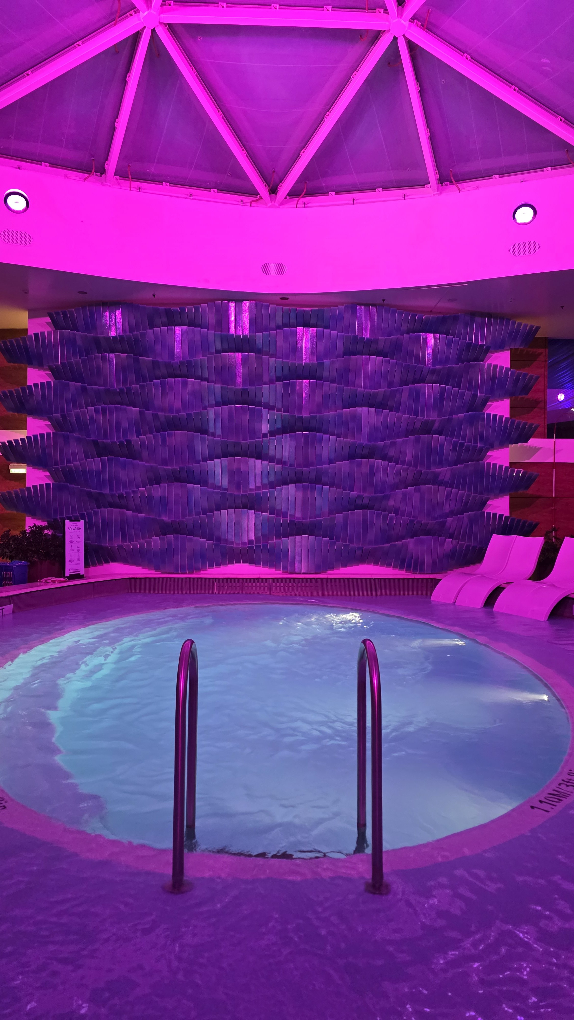 Indoor pool area illuminated with vibrant pink and purple lighting, featuring a decorative wall with a textured pattern and a set of white lounge chairs.