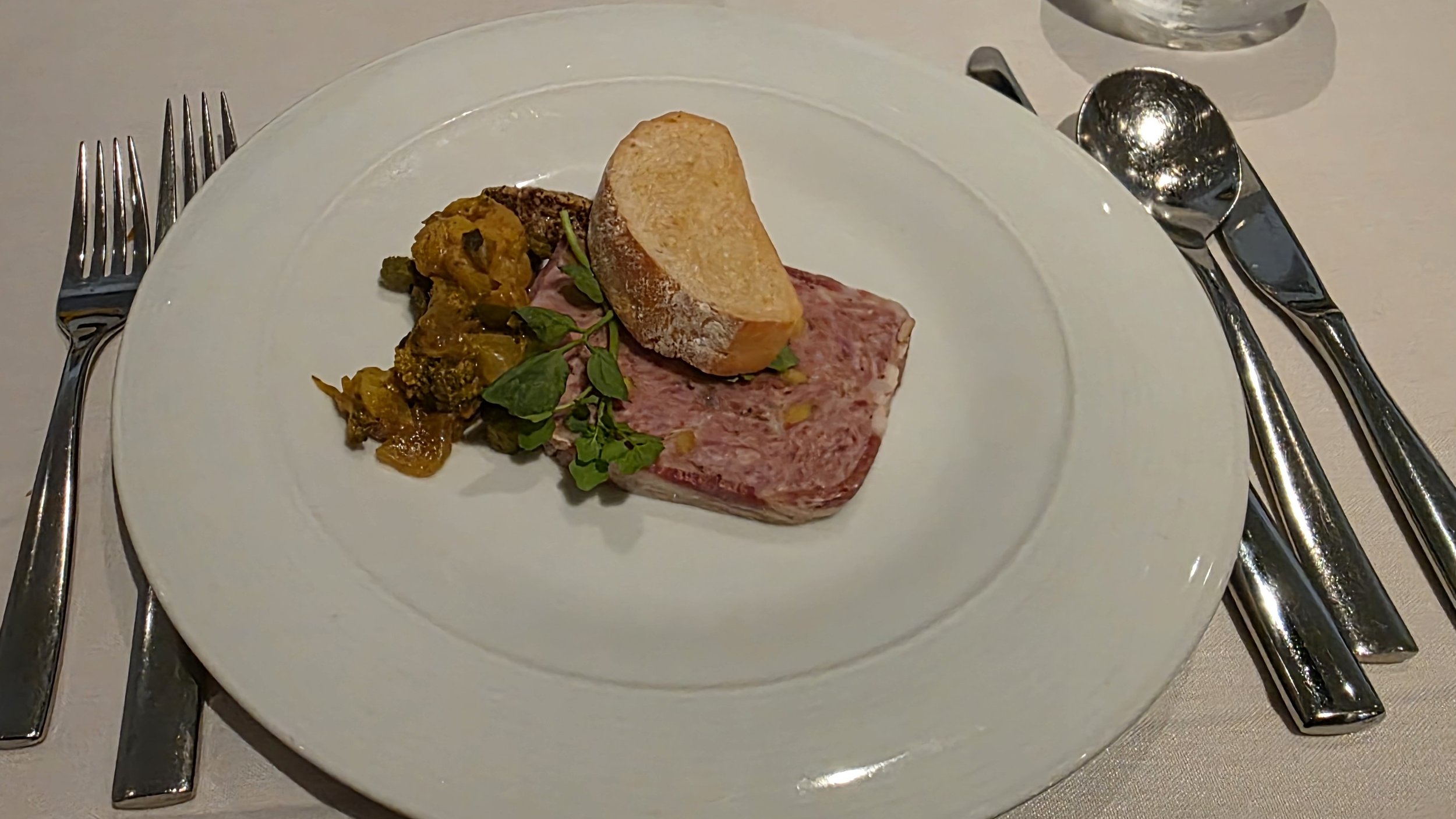 A fine dining plate with a slice of terrine, a raw beef patty with herbs, and a scoop of foie gras, garnished with a sprig of fresh herbs, on a white round plate.