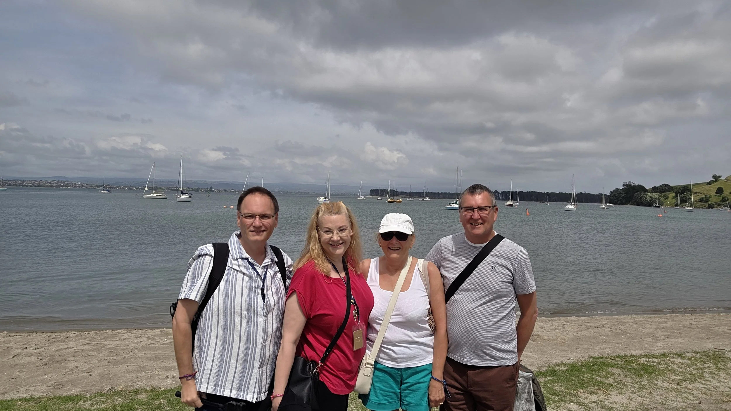 Group of four people standing on a beach near a body of water with sailboats, cloudy sky, and hills in the background.