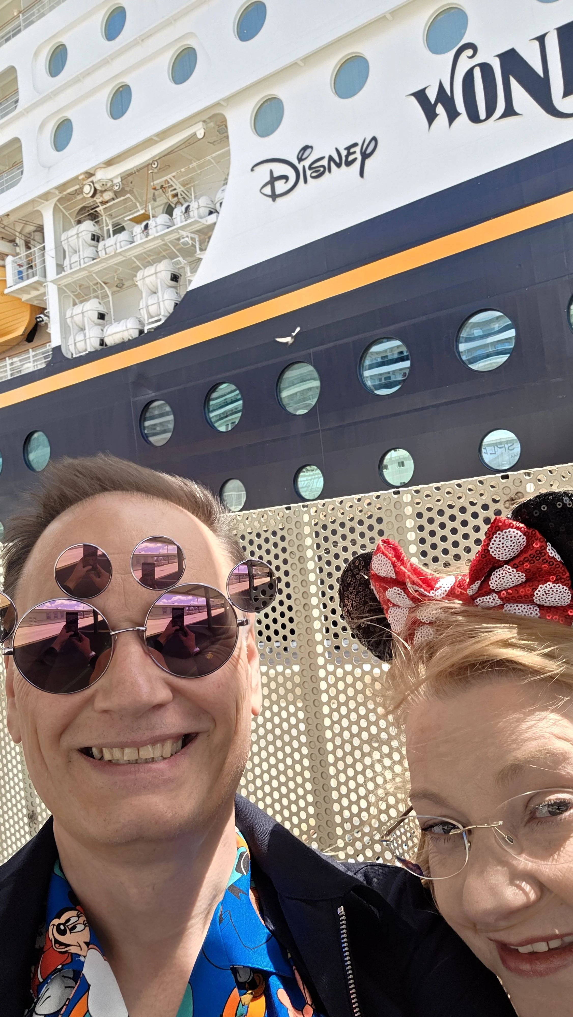 Two people smiling in front of a Disney cruise ship, one wearing round pink sunglasses and a Disney-themed shirt, the other wearing Minnie Mouse ears with a red and white polka dot bow.