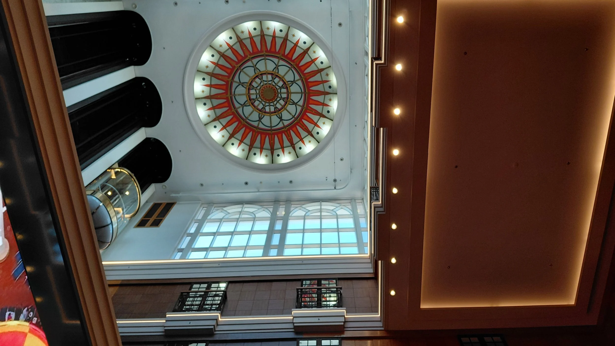 View of a hotel lobby looking upward at a decorative circular stained glass ceiling with red and gold sunburst design, surrounded by a skylight and walls with windows and balconies.