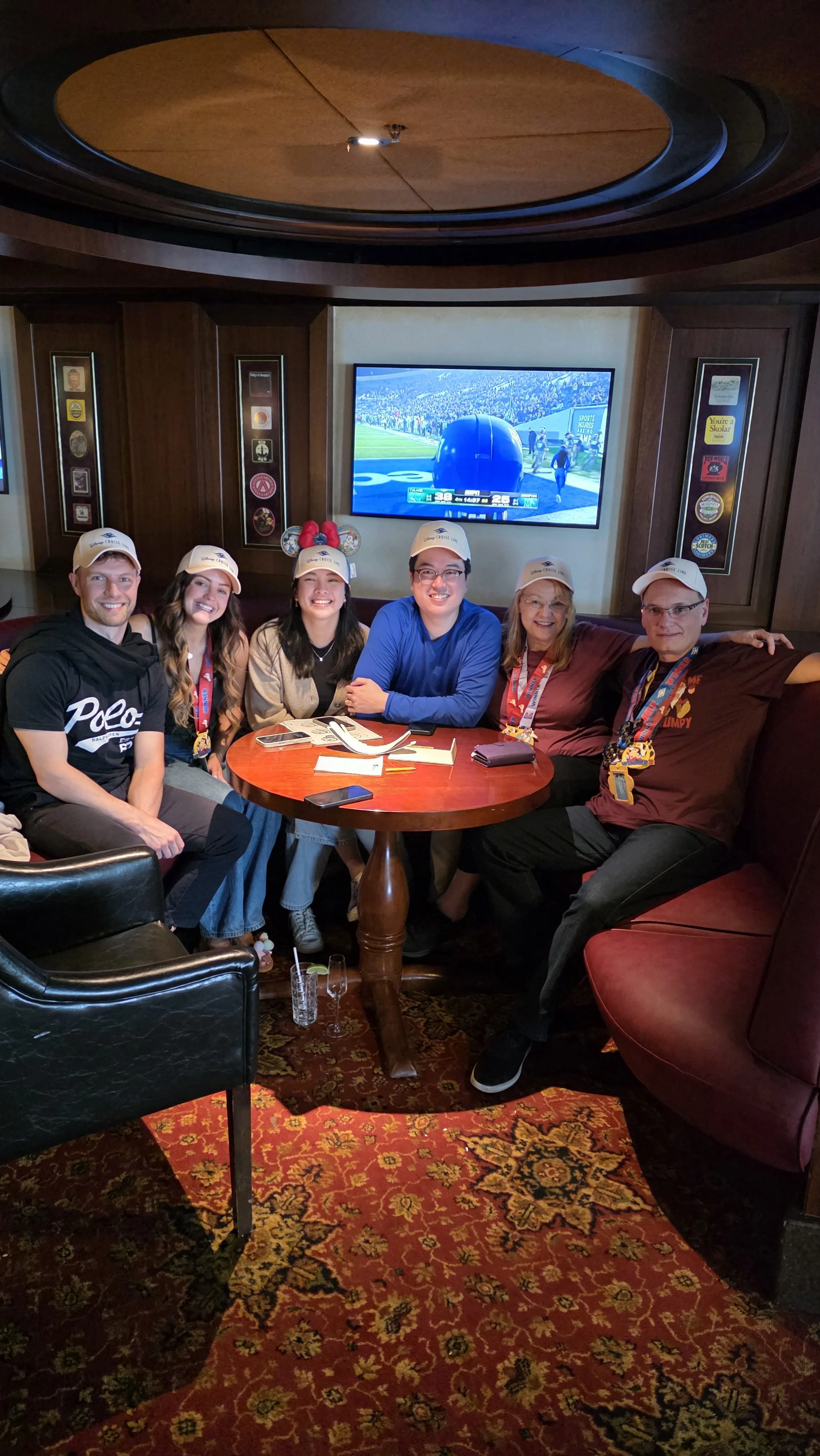 A group of six people sitting around a small round table in a restaurant, all wearing white baseball caps, smiling, and looking at the camera, with a TV showing a football game in the background.