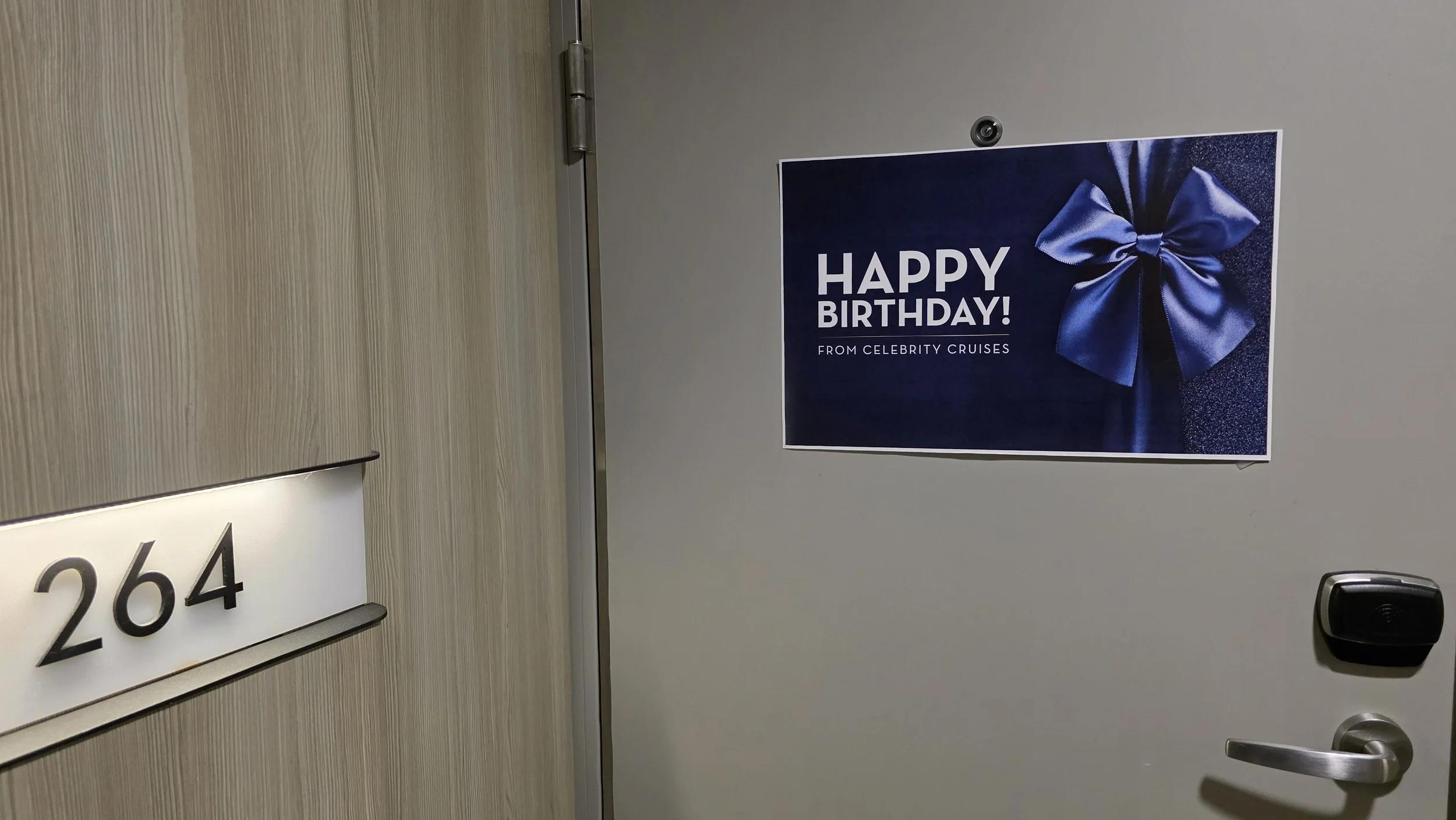 Door with room number 264 on a nameplate and a birthday sign with a blue ribbon bow that reads "Happy Birthday! From Celebrity Cruises".