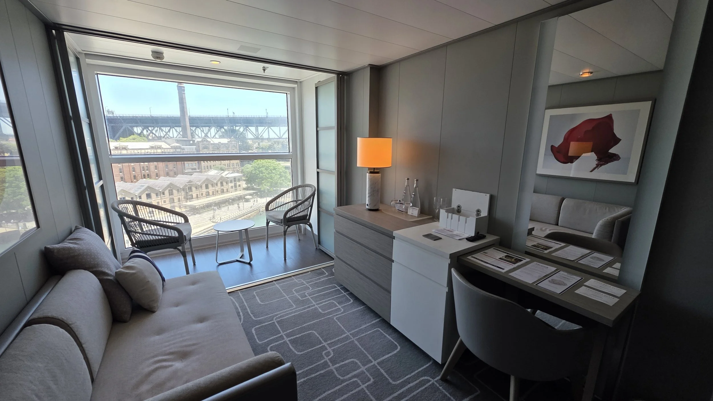 Modern hotel room with a large window overlooking city and bridge, beige sofa with pillows, small sitting area outside window, bedside table with lamp, and writing desk with brochures.