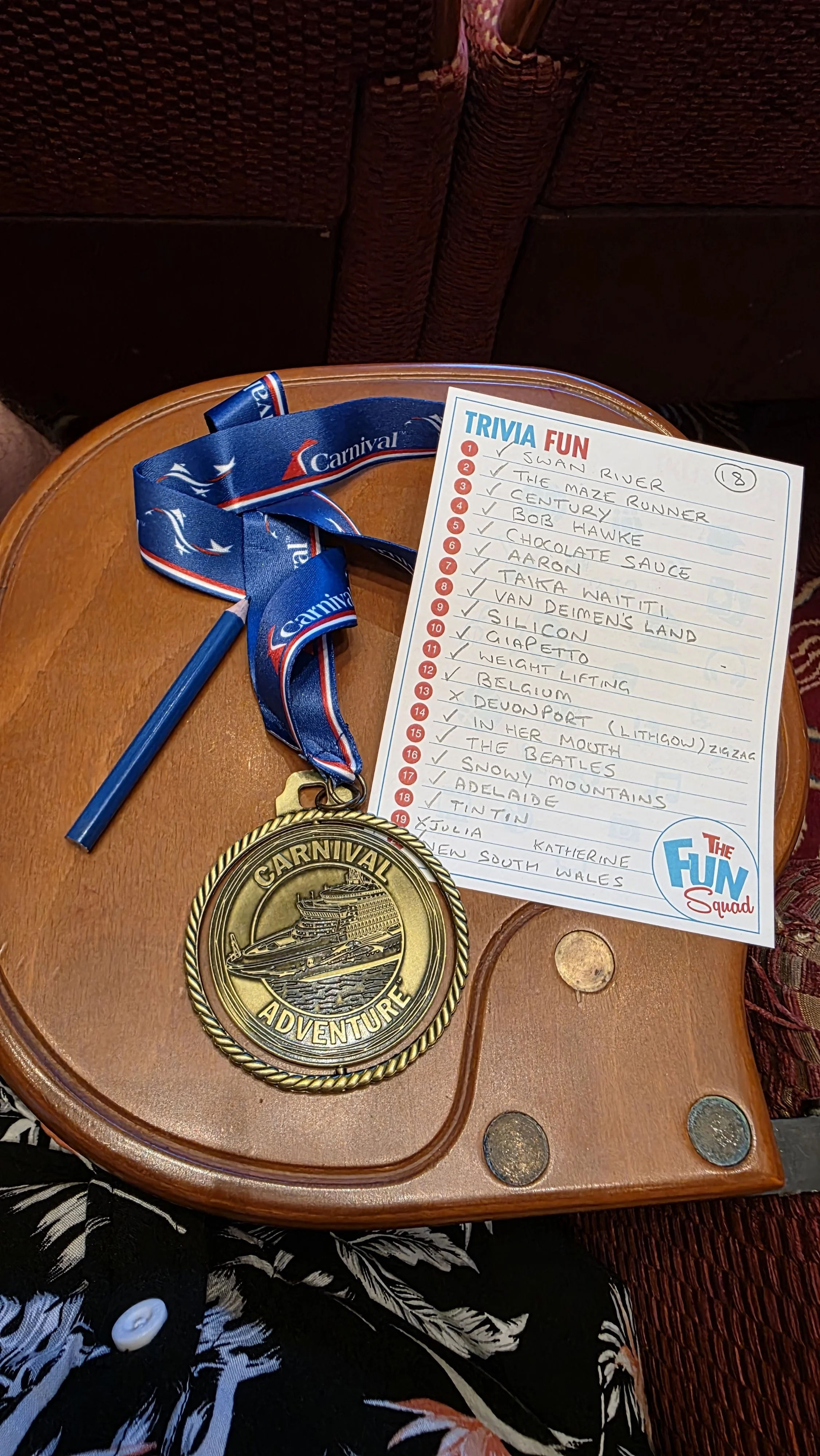 A gold-colored Carnival adventure medal with a cruise ship engraved on it, attached to a blue ribbon with 'Carnival' branding, resting on a round wooden table. Next to the medal is a trivia fun sheet with a list of 20 questions, most of which are che