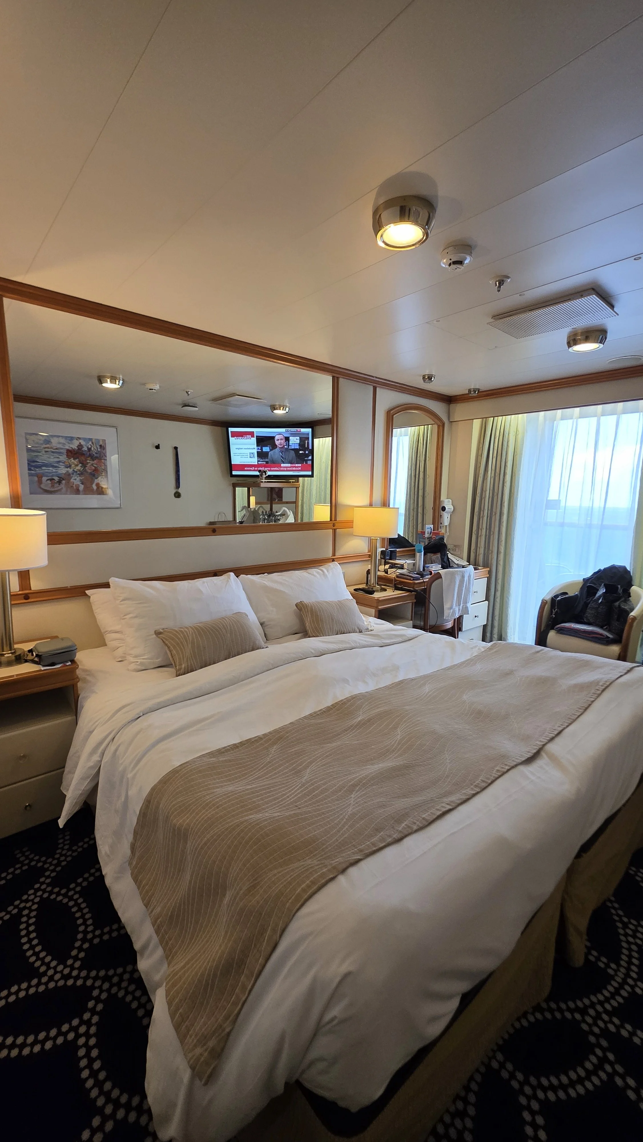 A hotel room with a large bed, beige bedspread, and multiple pillows, side tables with lamps, a chair with a backpack, a TV on the wall, and a large window with curtains.