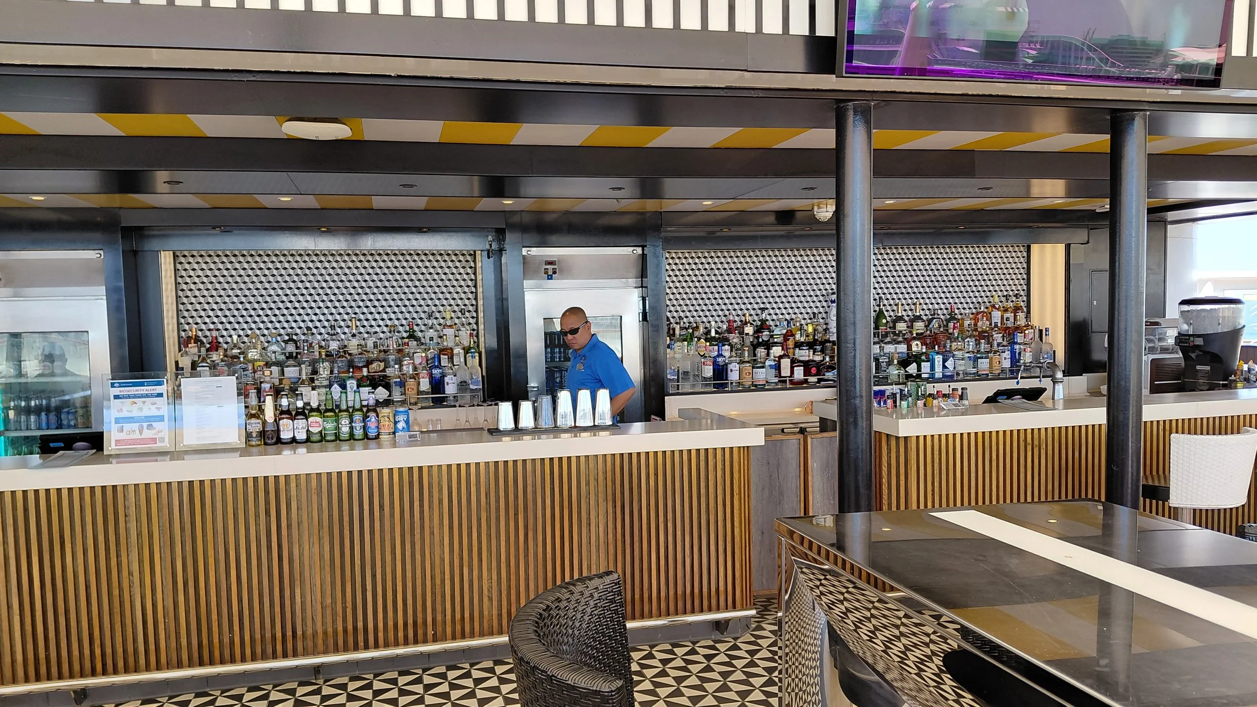 A bartender wearing a blue shirt and sunglasses standing behind a bar counter filled with various bottles of alcohol, glasses, and bar equipment. The background features a textured metallic wall, with a portion of a television screen visible at the t
