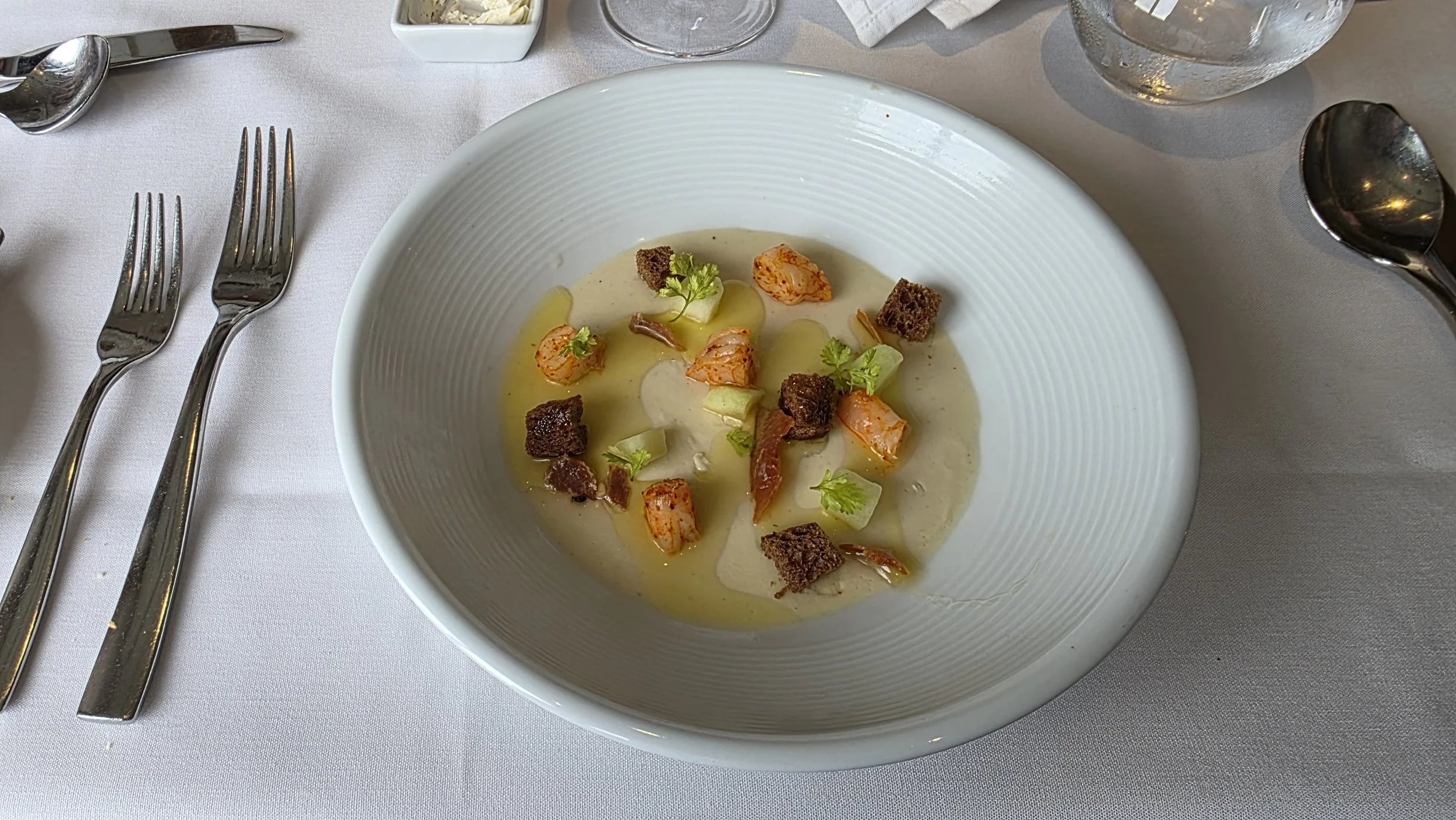 A bowl of creamy soup garnished with chopped herbs, diced vegetables, bacon bits, croutons, and a drizzle of oil on a white tablecloth, with cutlery and glassware nearby.