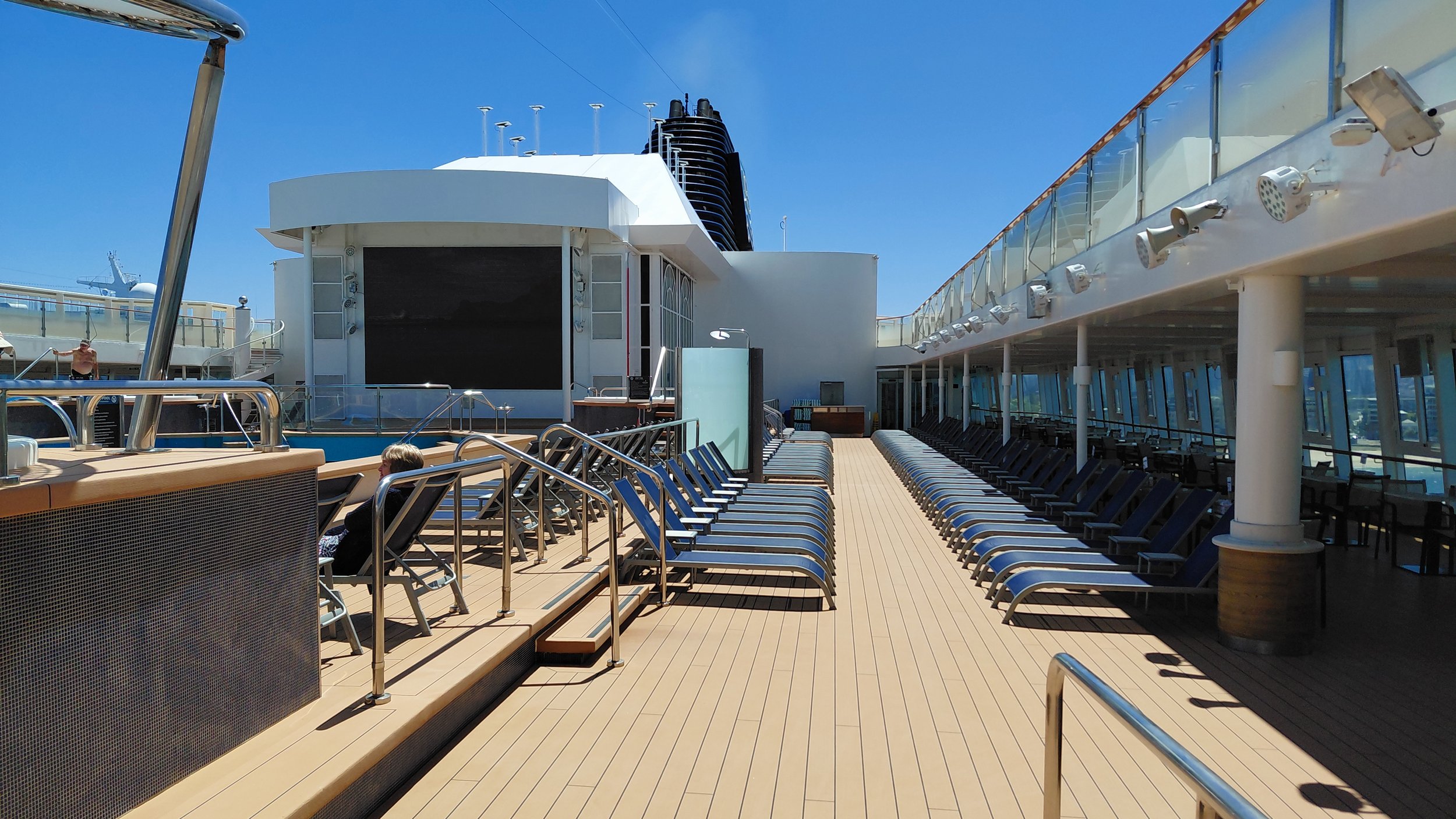 Empty pool deck on a cruise ship with lounge chairs, a large outdoor movie screen, and glass railings under a clear blue sky.