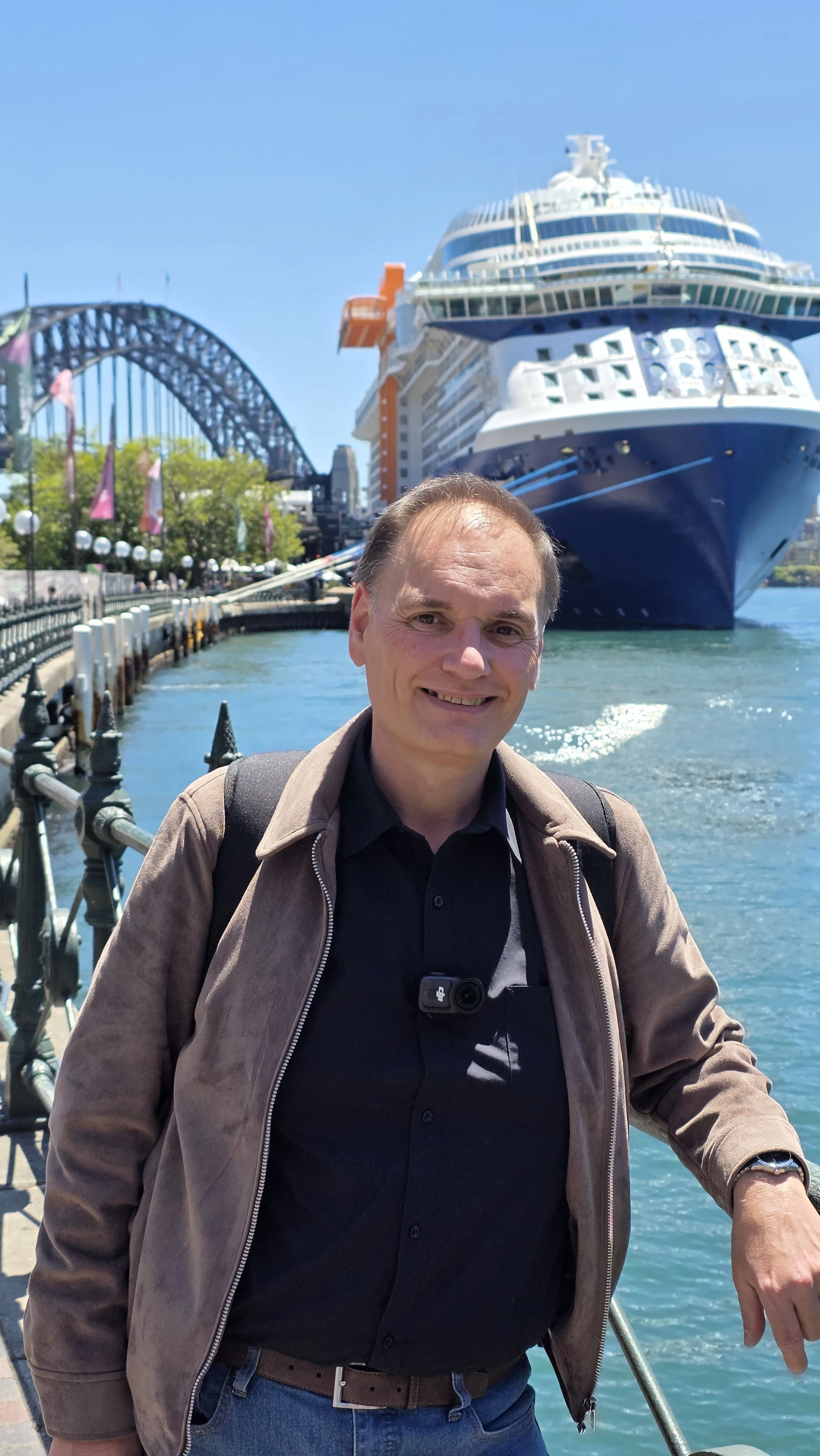 A man stands on a waterfront promenade with a large cruise ship behind him and a bridge in the distance on a clear day.