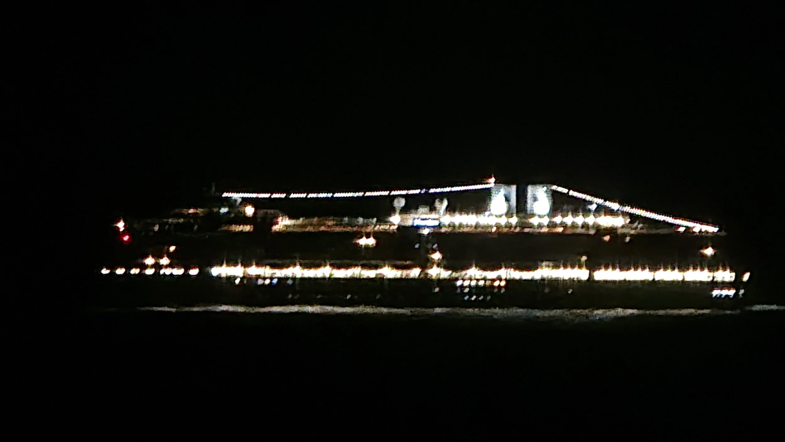 The image shows a large, dark vehicle illuminated with bright lights along its edges and surface, likely a bus or a similar type of transportation, against a black background at night.