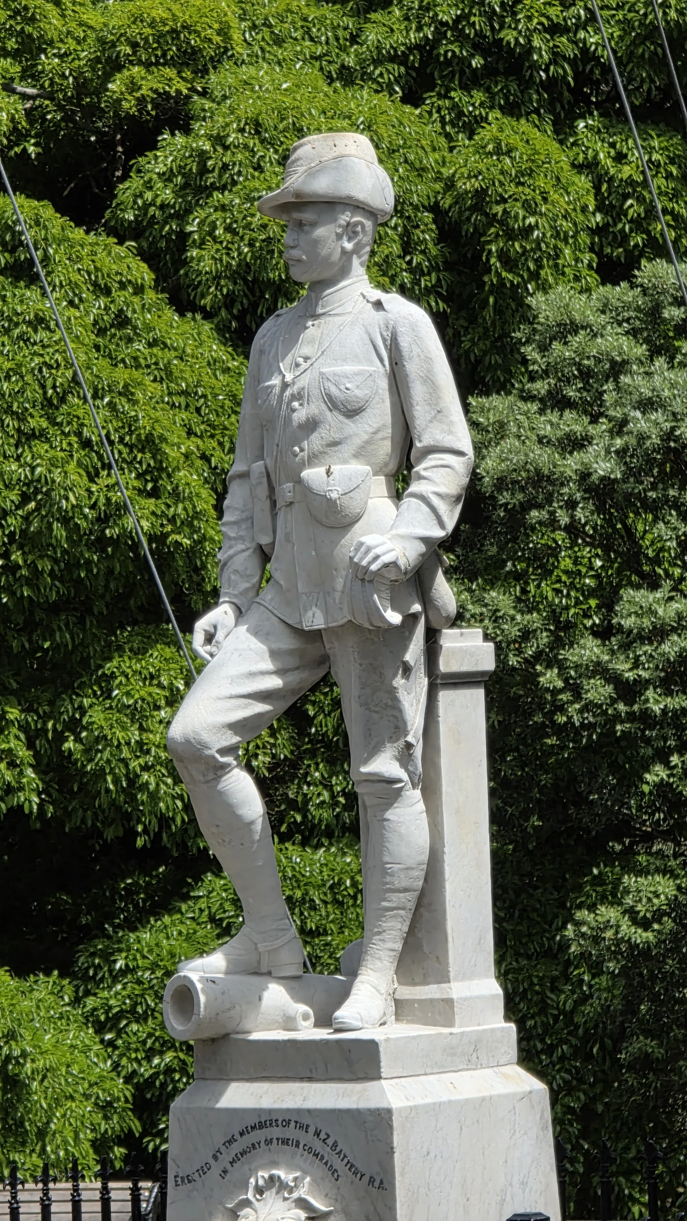 A white statue of a soldier in uniform with a hat, standing with one foot on a cannon, set against green foliage.