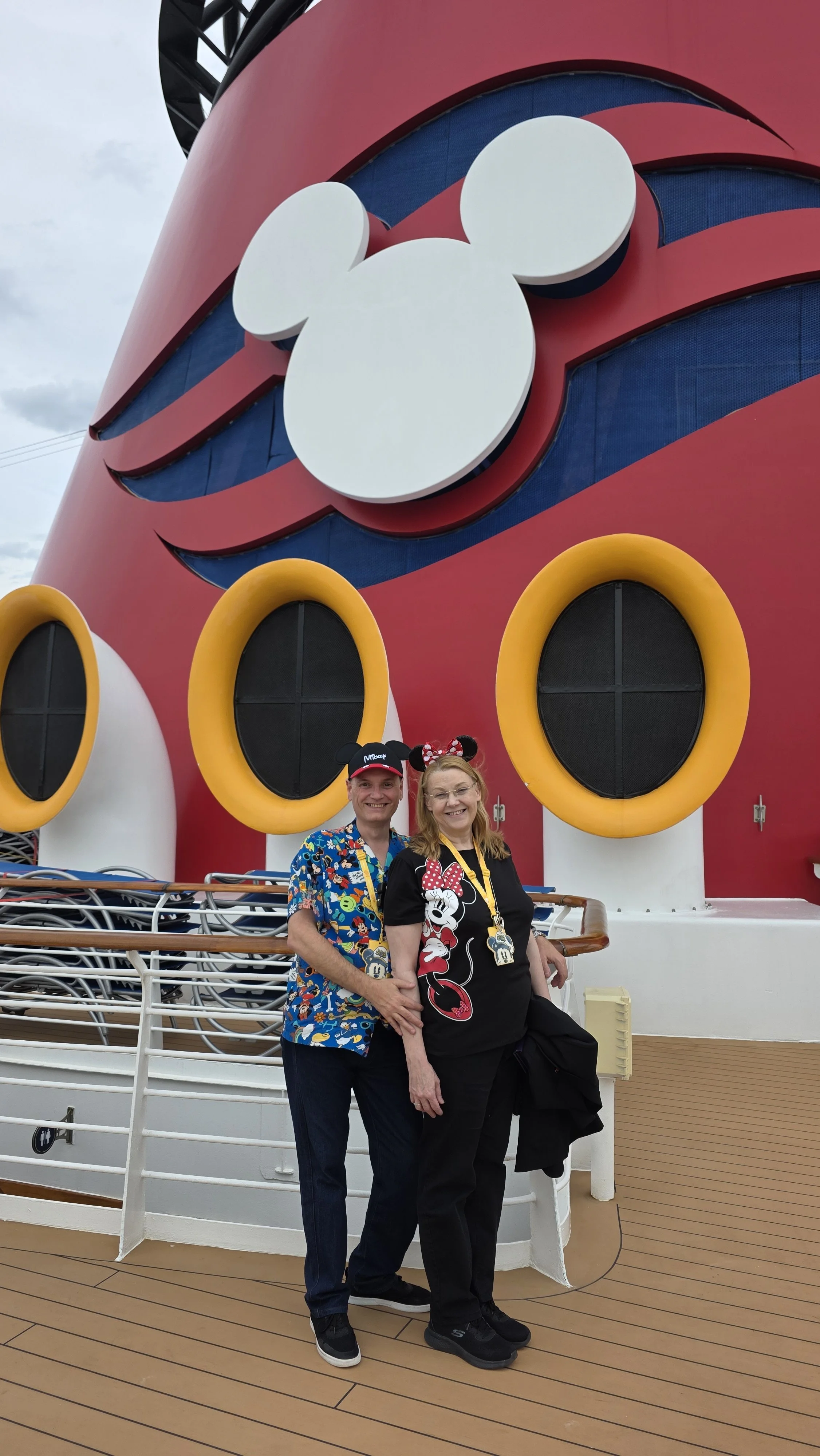 Two people standing in front of a large cruise ship with Minnie Mouse themed decorations. The woman is wearing Minnie Mouse ears and a black shirt with Minnie Mouse on it, and the man is wearing a colorful Disney-themed shirt and a Disney cap.