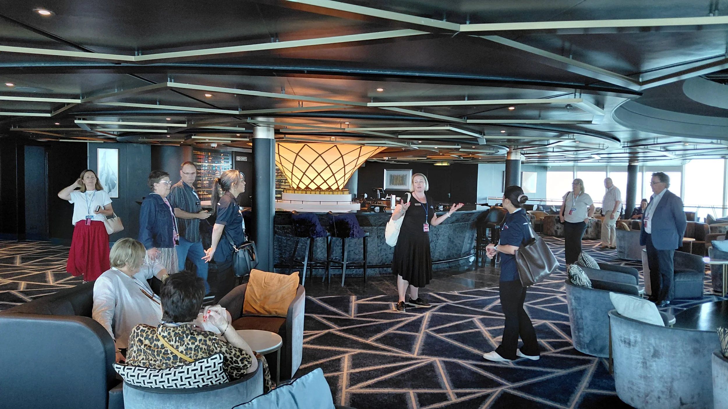 A group of people congregate in a modern, upscale lounge with a patterned carpet and stylish furniture, with a woman in black explaining something to a tour group, some seated and some standing, with large windows in the background.