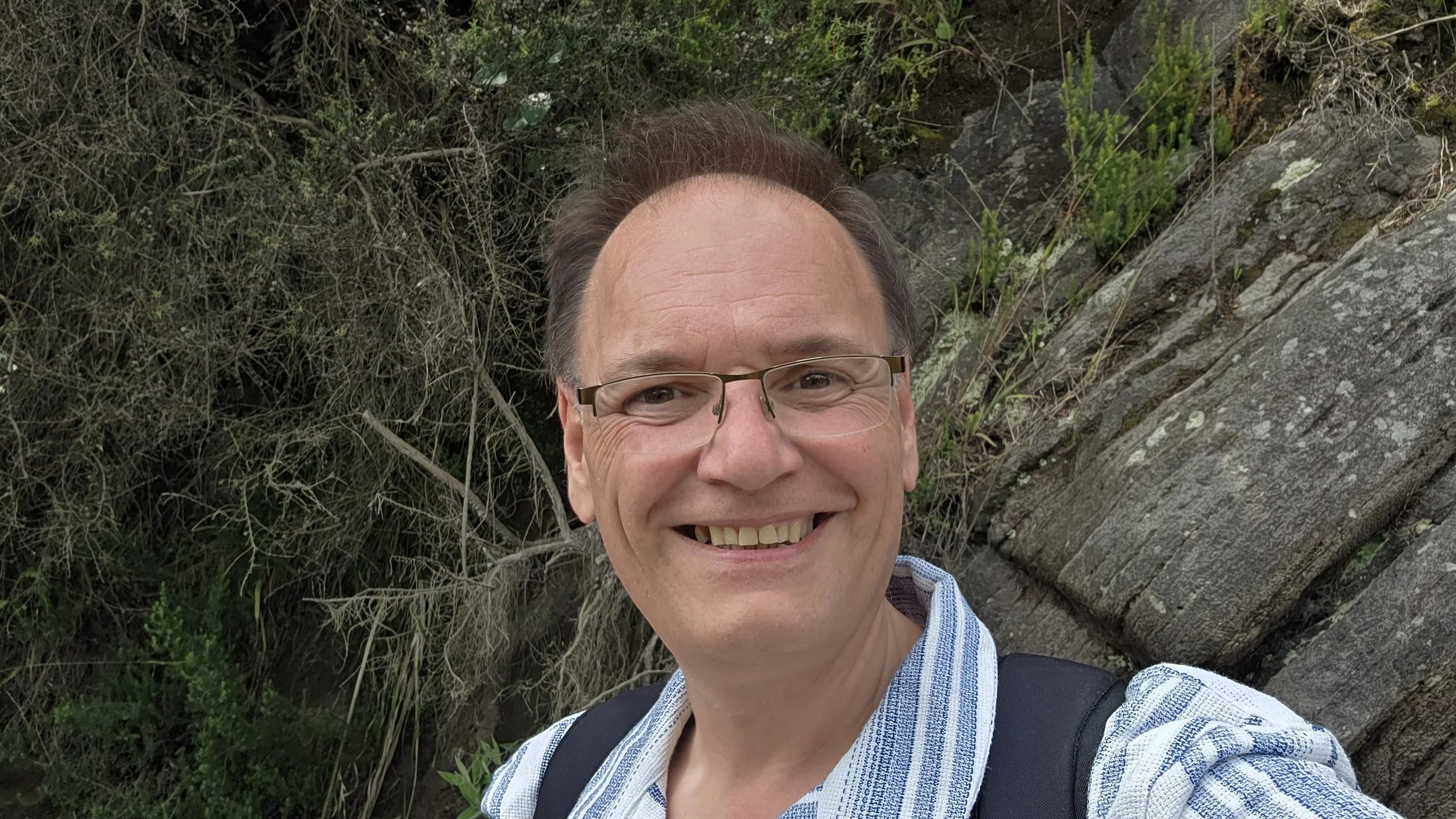 A man with glasses, smiling outdoors, wearing a striped shirt and backpack, with trees and rocks in the background.