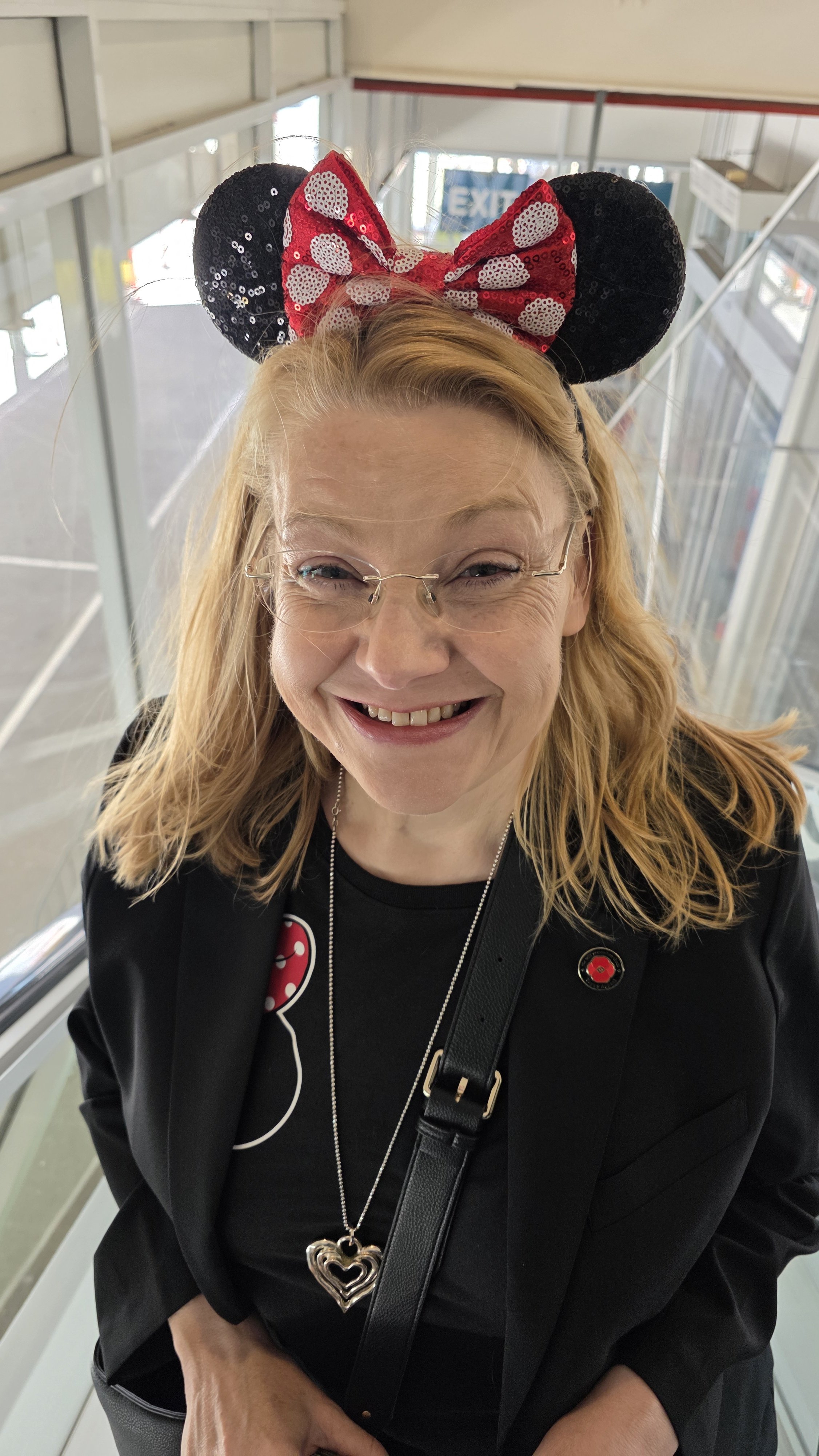A smiling woman wearing glasses, a Minnie Mouse headband, and a black shirt with badges, standing inside an airport terminal near a glass window.