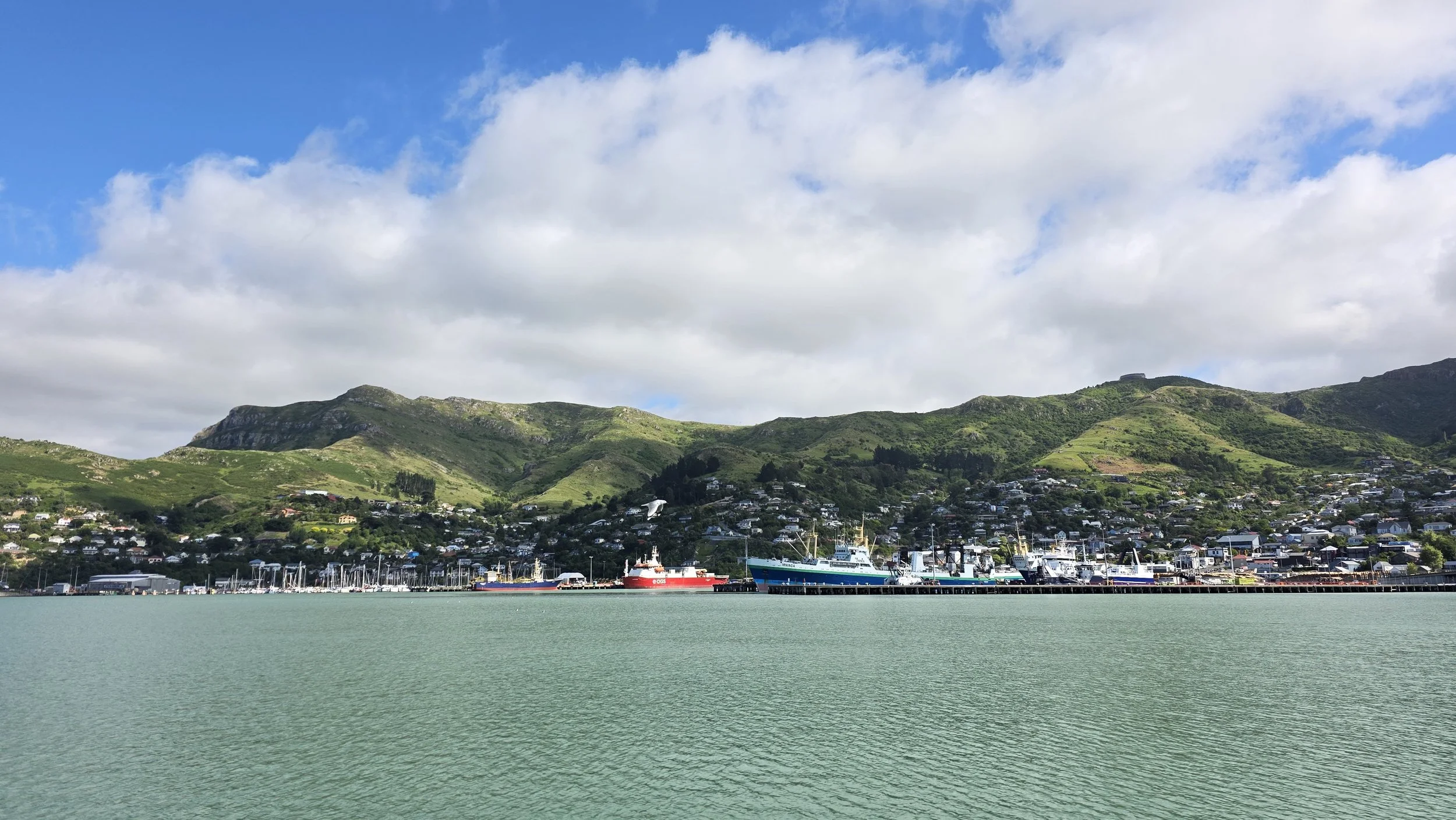 Harbor with boats docked, green hills in the background, and partly cloudy sky.