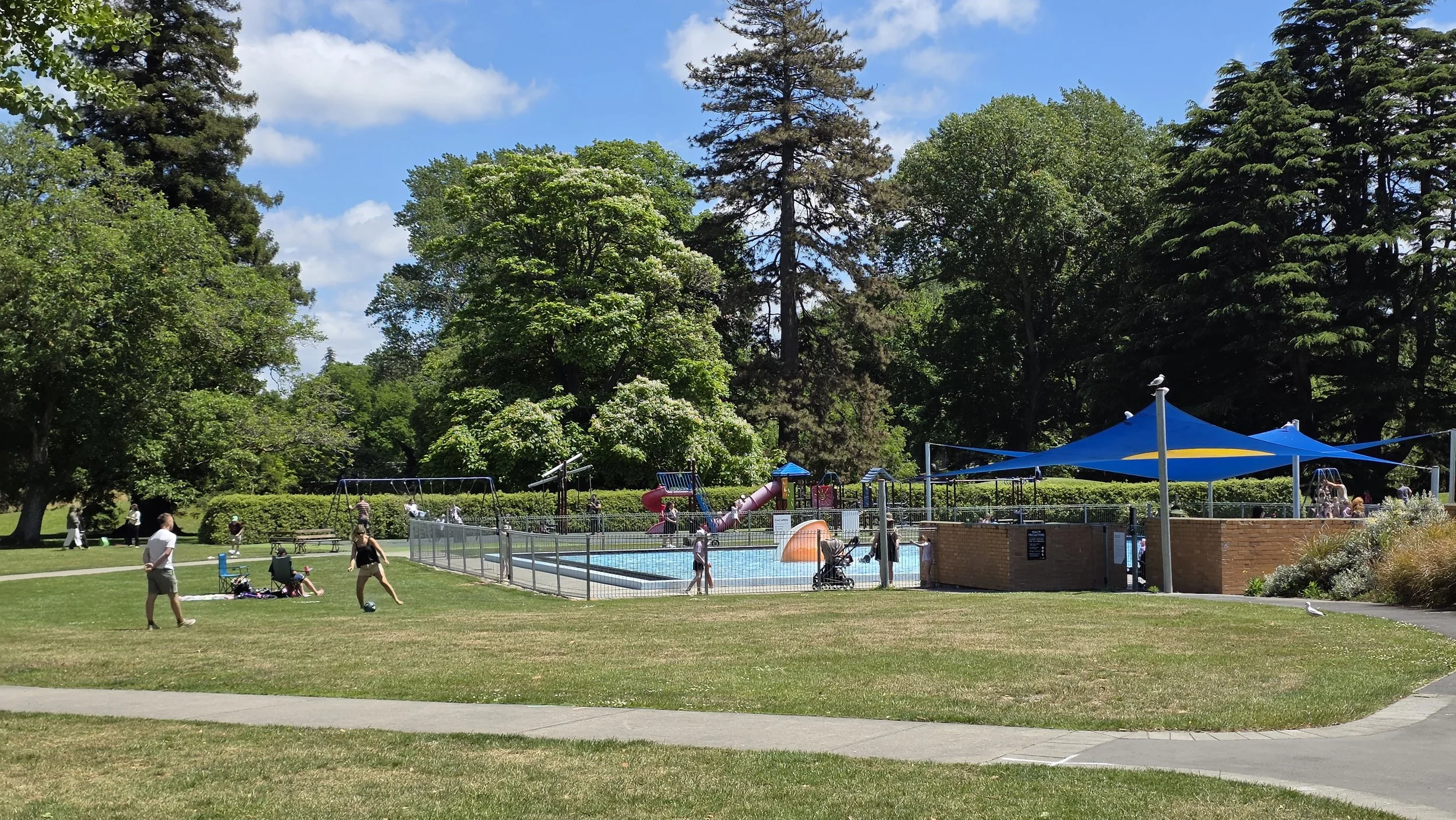 Public park with a swimming pool, colorful water slide, shaded area with blue canopy, children playing, people walking, and large green trees under a partly cloudy sky.