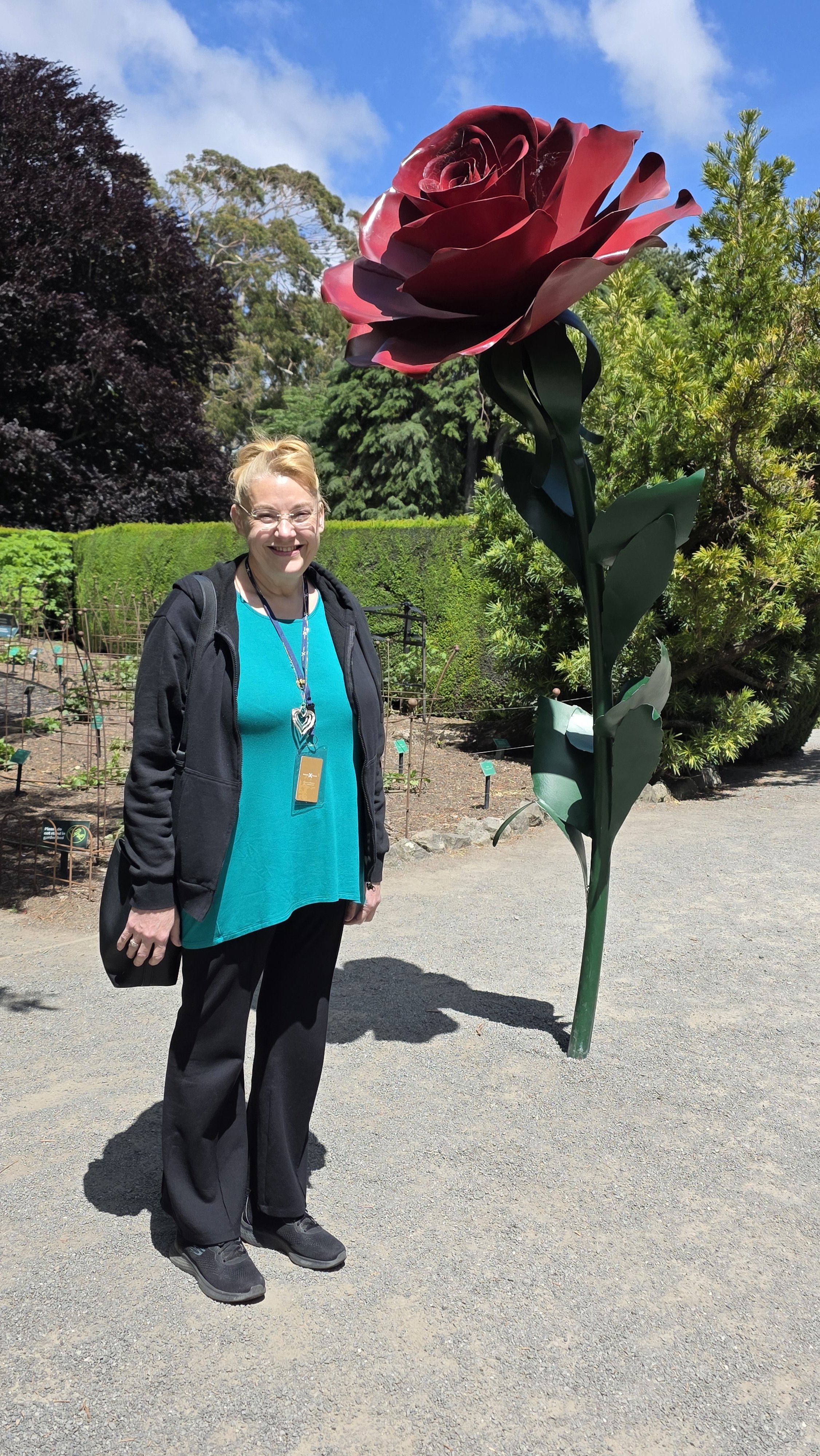 A smiling woman wearing glasses, a black jacket, teal top, black pants, and black shoes standing next to a large artificial red flower sculpture outside in a garden or park with trees and a hedge in the background.