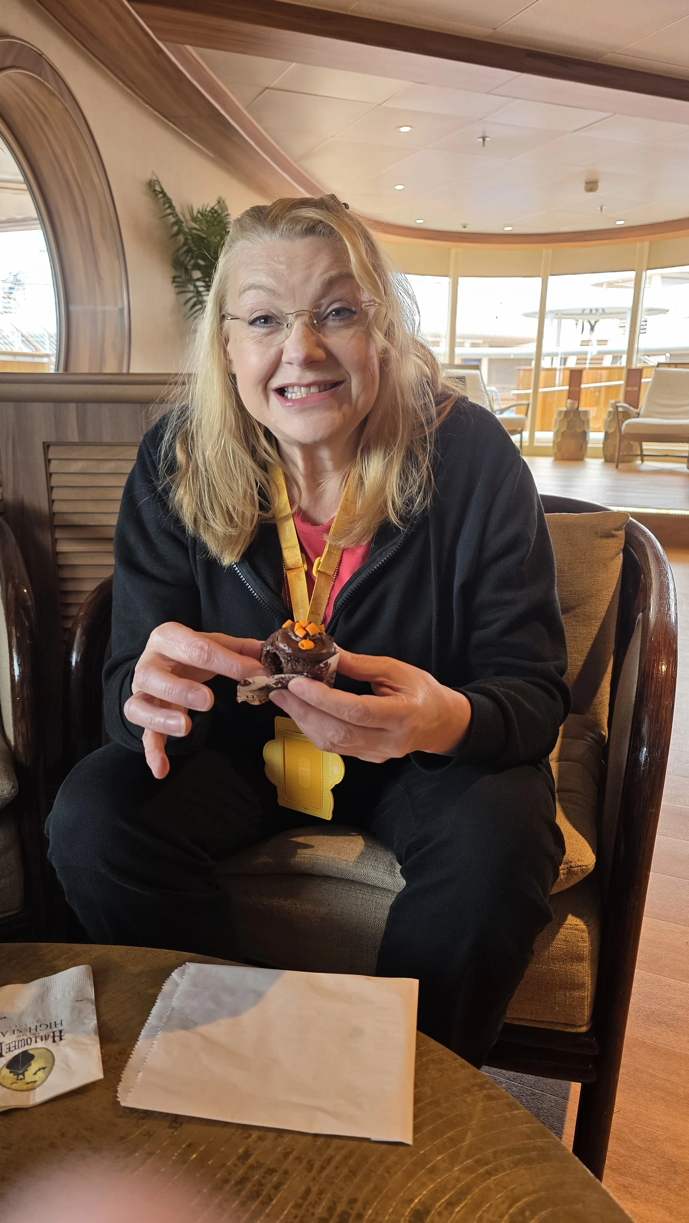 A woman with blonde hair, glasses, and a black jacket sitting on a chair in a wooden interior, holding a cupcake decorated with orange icing, smiling at the camera.