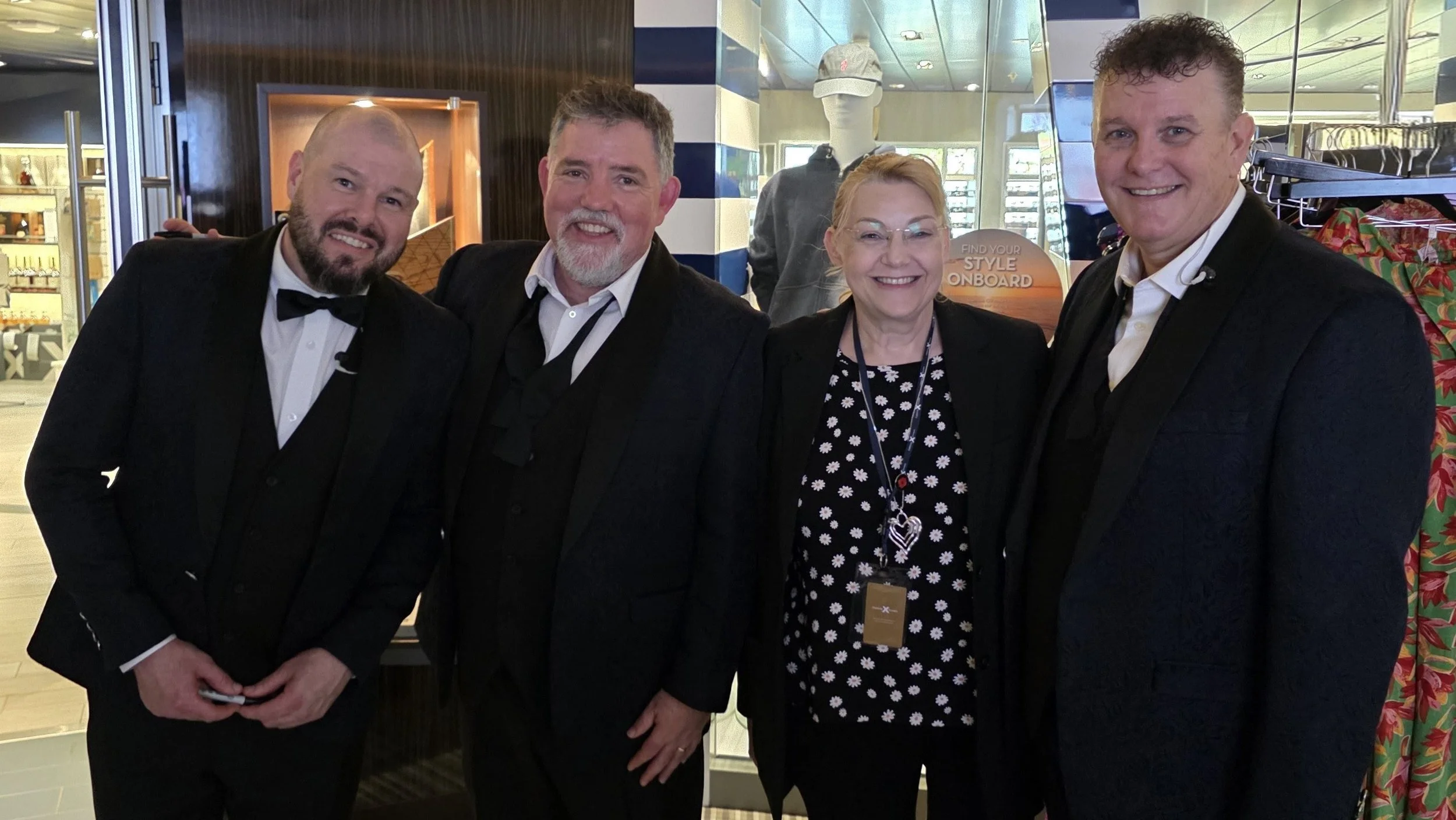 Four people dressed in formal attire standing close together and smiling indoors, with some store displays in the background.