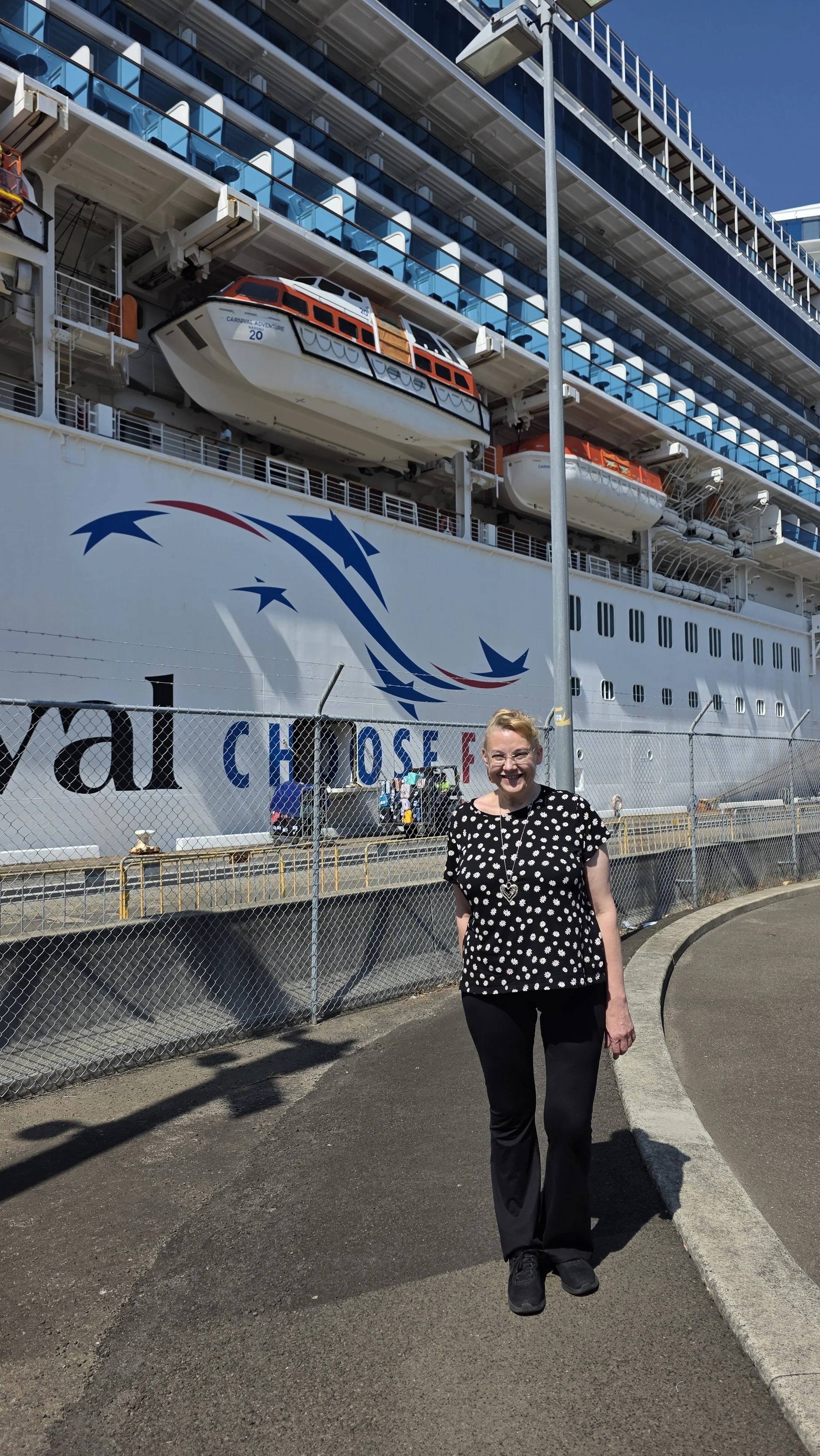 A woman standing on a sidewalk in front of a large cruise ship with lifeboats and a white hull. The woman is smiling, wearing a black shirt with white polka dots and black pants.