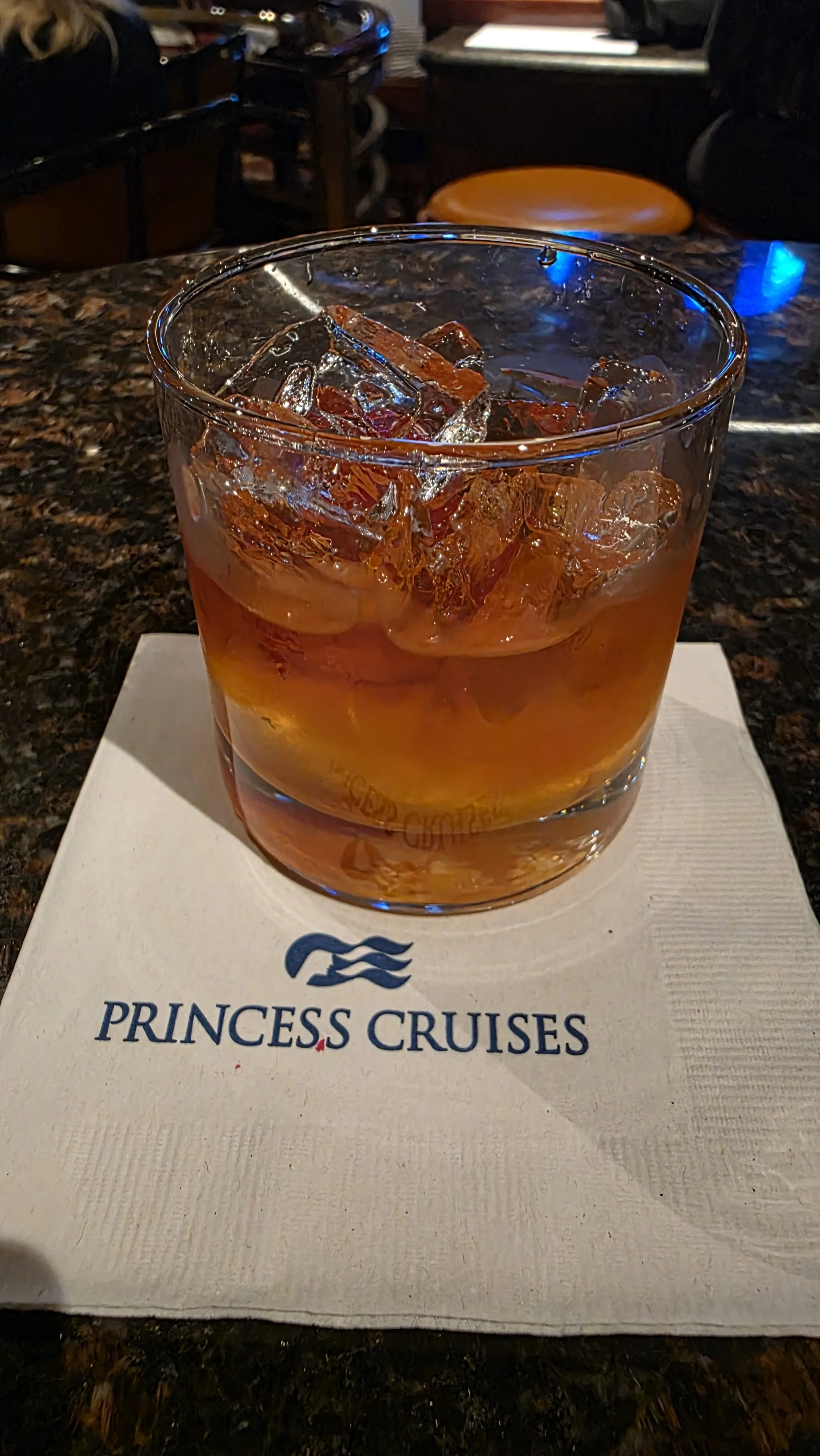 A glass of amber-colored beverage with ice cubes on a napkin that has the Princess Cruises logo, on a dark marble bar counter.