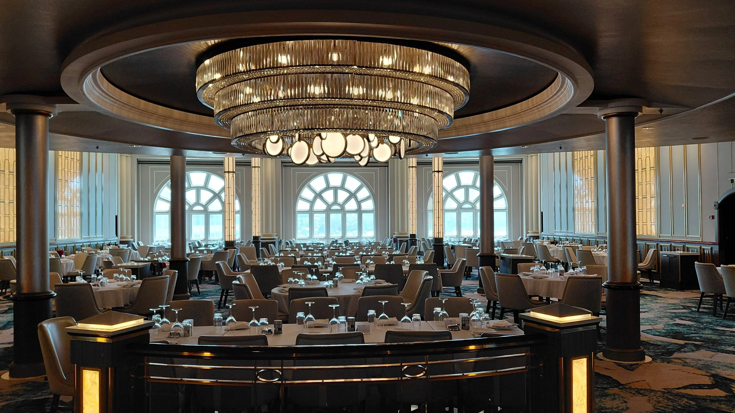 Elegant banquet hall with round tables set for dining, large windows, a grand chandelier hanging from a circular ceiling, and ornate wall panels.