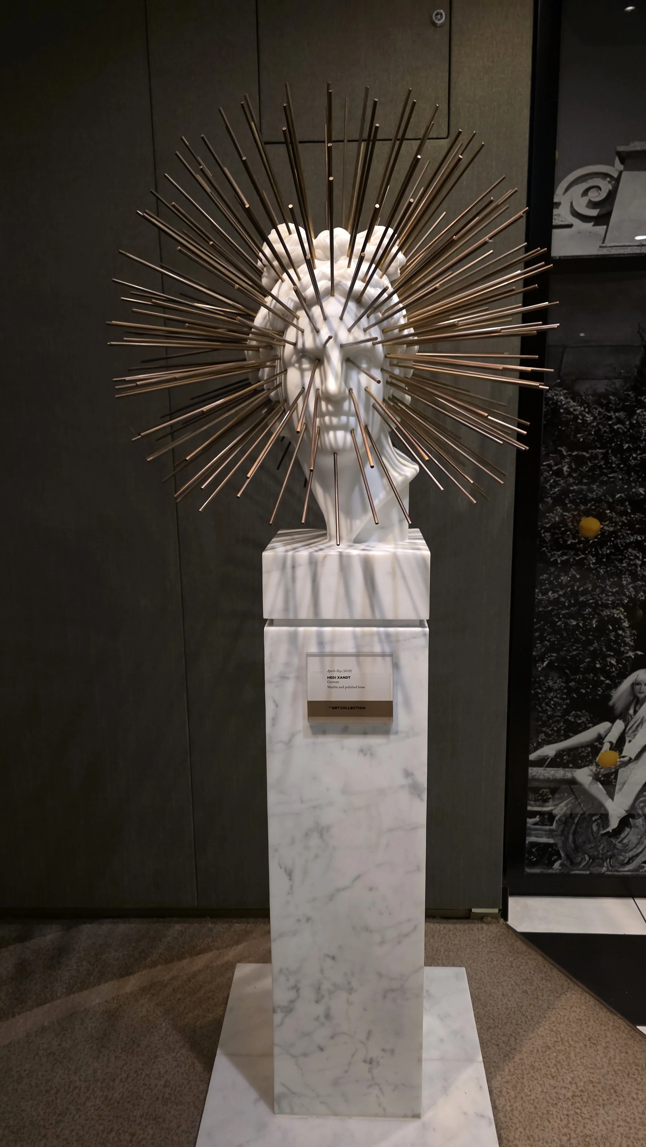 Modern sculpture of a human face with a halo of metal rods, displayed on a marble pedestal in an art gallery.