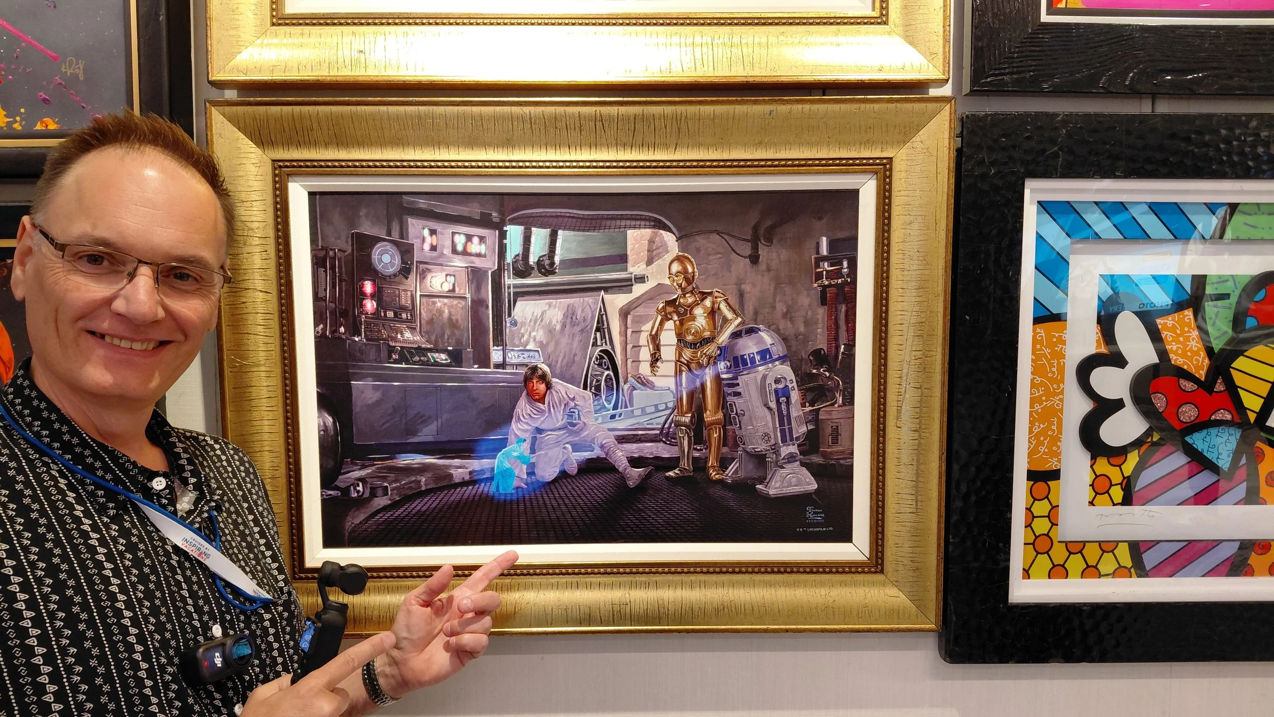 A man in glasses smiling and pointing at a framed 'Star Wars' artwork on a gallery wall.