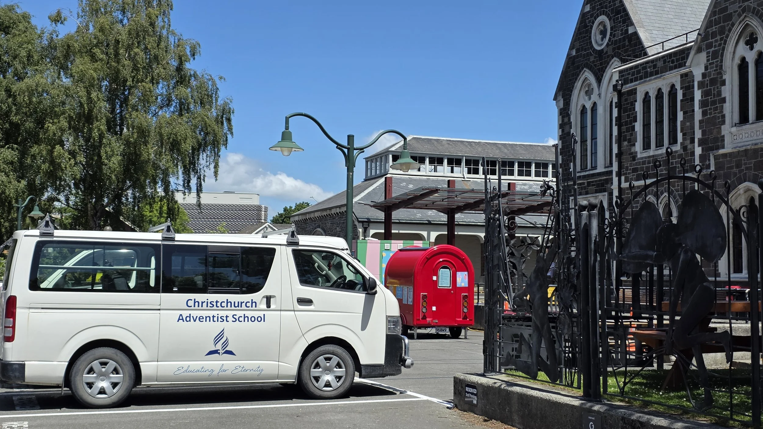 White van with blue lettering that reads 'Christchurch Adventist School' parked in front of a church with black stone walls and stained glass windows, a red mailbox, an iron gate with religious figures, and a leafy tree under a bright blue sky.