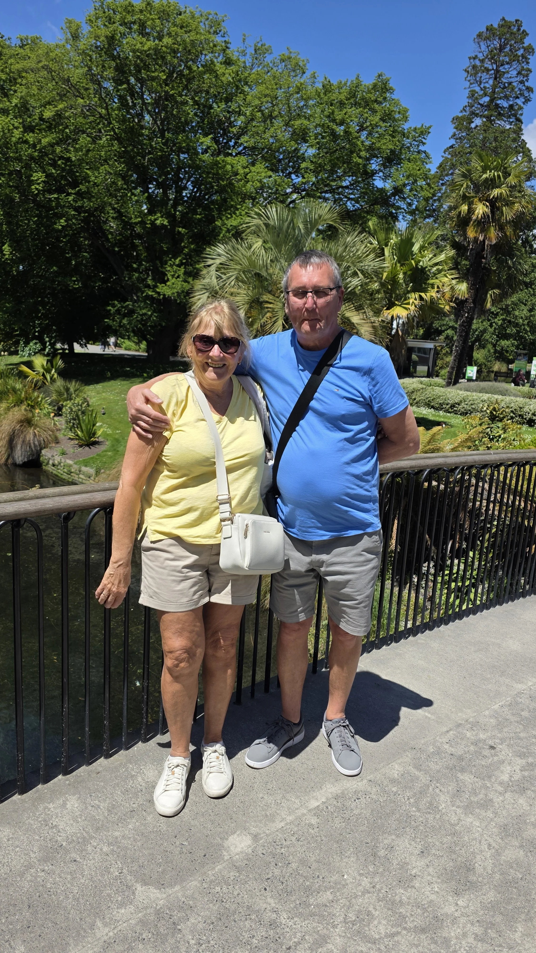 A smiling elderly woman and a man standing side by side on a bridge in a park with green trees and palm trees in the background on a sunny day.